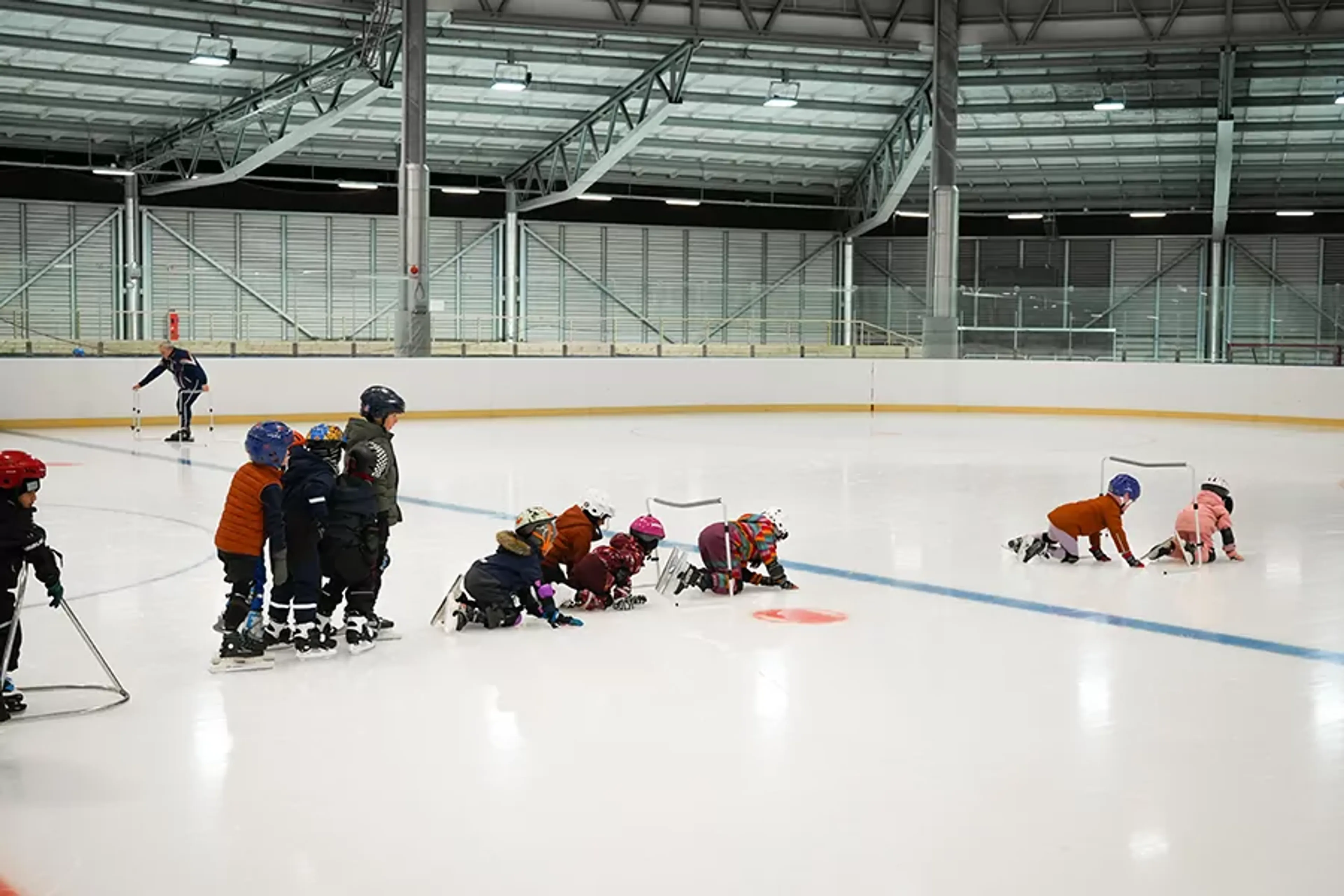 Barn i hjälmar och vinterkläder övar på att åka skridskor på en inomhusrink med hjälp av stöd och handledning. En ledare syns i bakgrunden som följer aktiviteten.