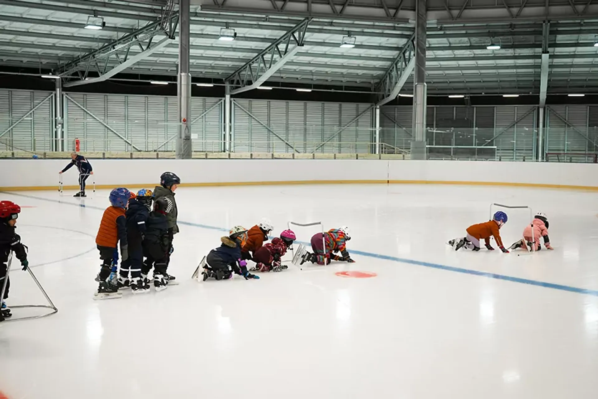 Barn i hjälmar och vinterkläder övar på att åka skridskor på en inomhusrink med hjälp av stöd och handledning. En ledare syns i bakgrunden som följer aktiviteten.