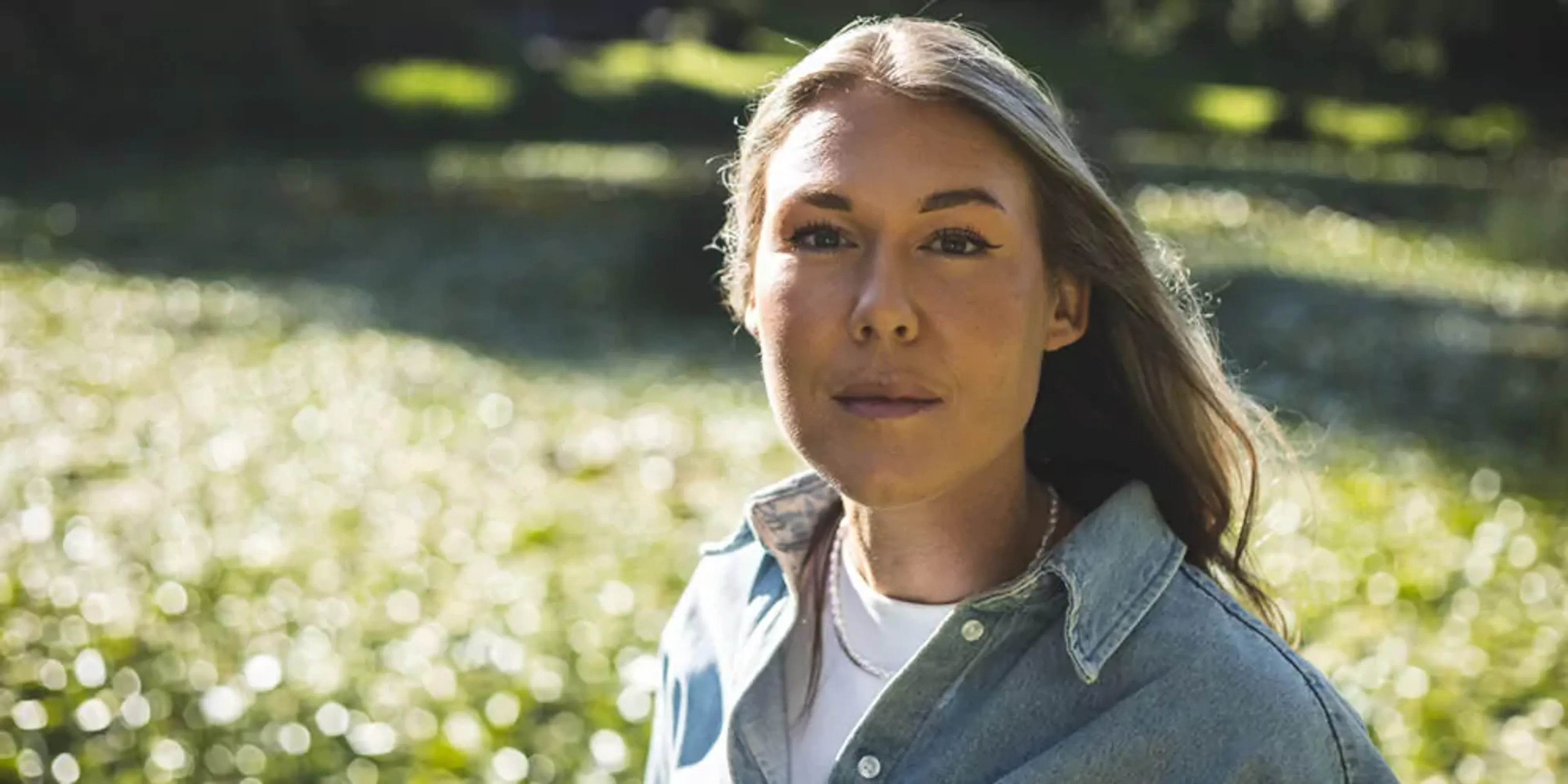 En kvinna med långt blondt hår och ljusblå skjorta står utomhus med gröna, suddiga bakgrundsdetaljer. Ljuset skapar ett mjukt skimmer runt henne.
