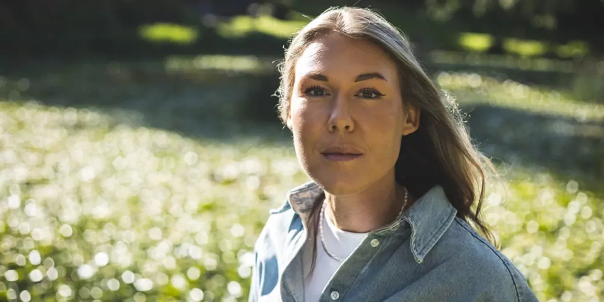 En kvinna med långt blondt hår och ljusblå skjorta står utomhus med gröna, suddiga bakgrundsdetaljer. Ljuset skapar ett mjukt skimmer runt henne.