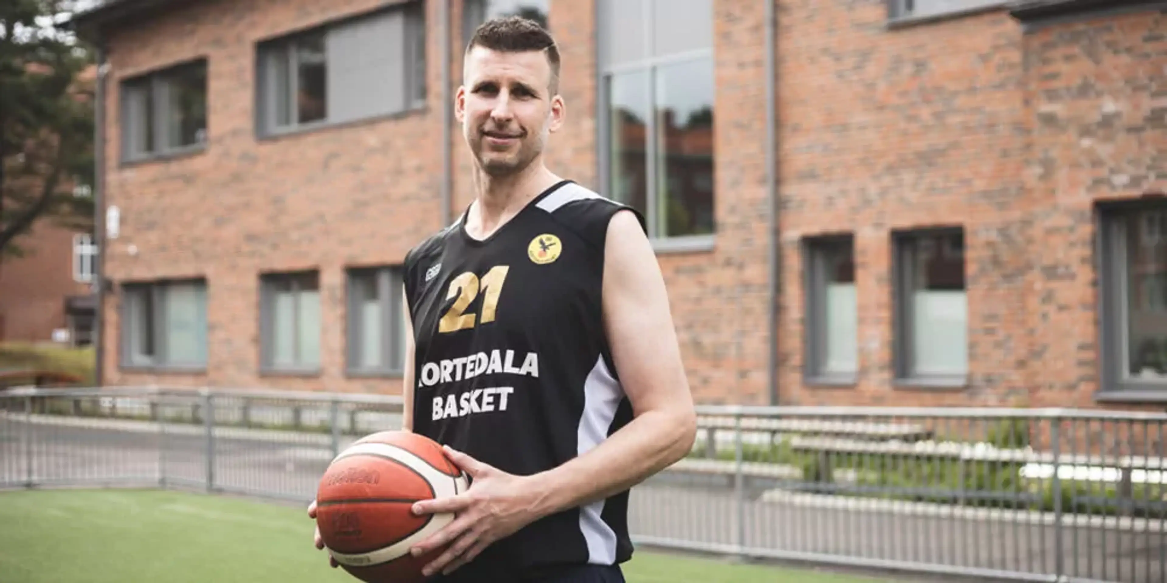 En man i svart och vit baskettröja med nummer 21 står utomhus och håller en basketboll. Bakom honom syns en tegelbyggnad med flera fönster.