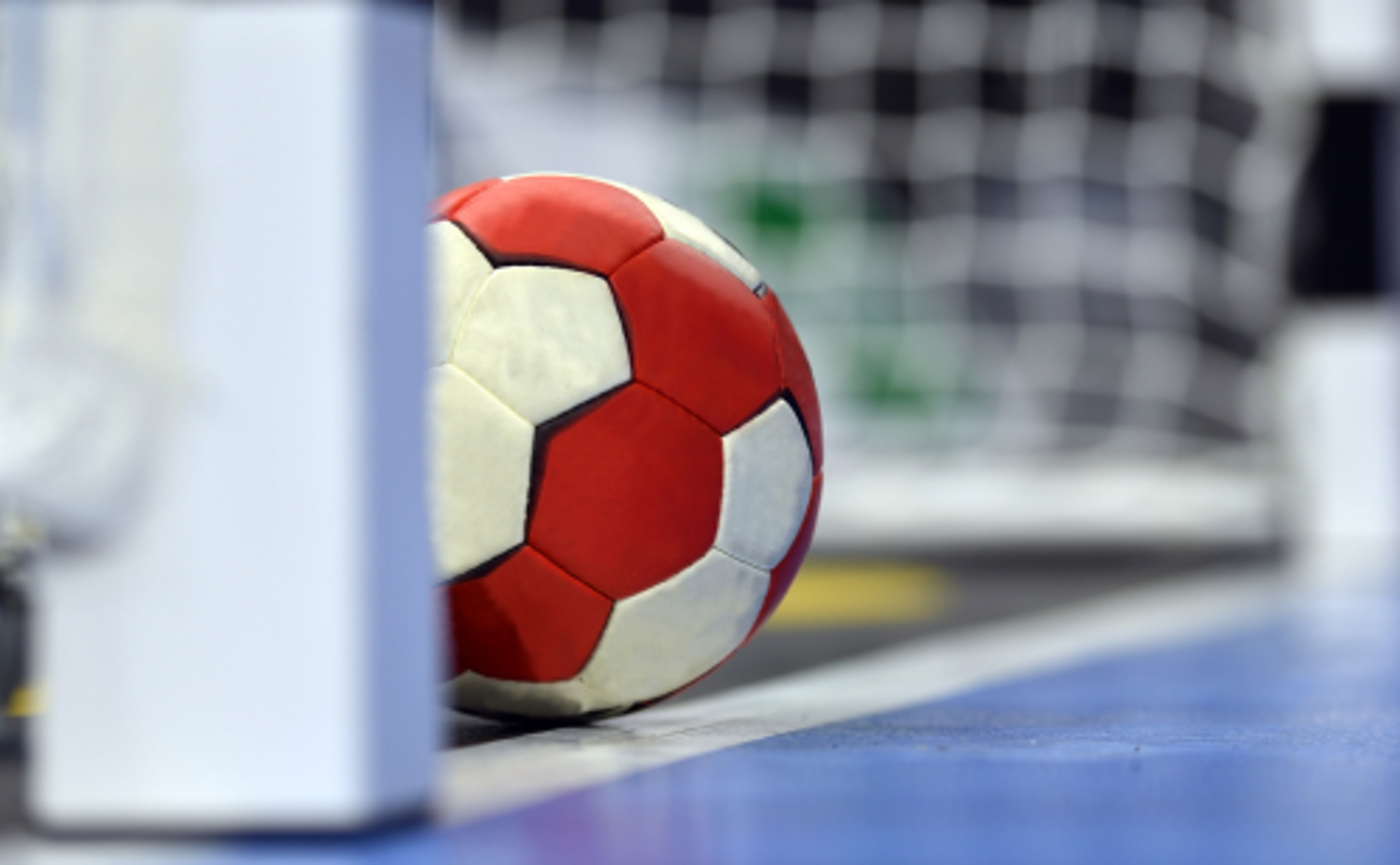 En röd- och vitmönstrad handboll som ligger vid mållinjen framför ett handbollsmål. Golvet är blått och bakgrunden suddig med nätet tydligt synligt.