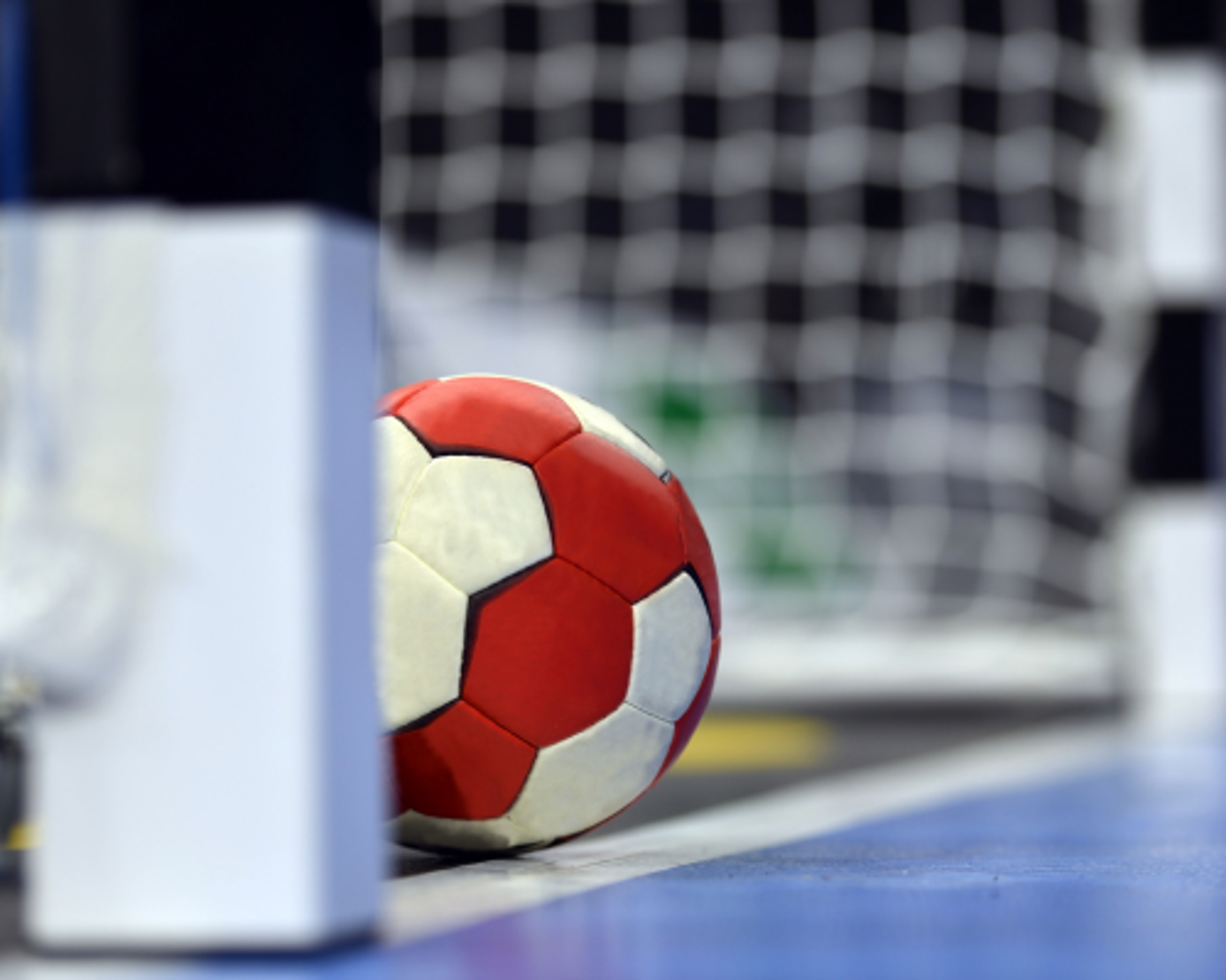 En röd- och vitmönstrad handboll som ligger vid mållinjen framför ett handbollsmål. Golvet är blått och bakgrunden suddig med nätet tydligt synligt.