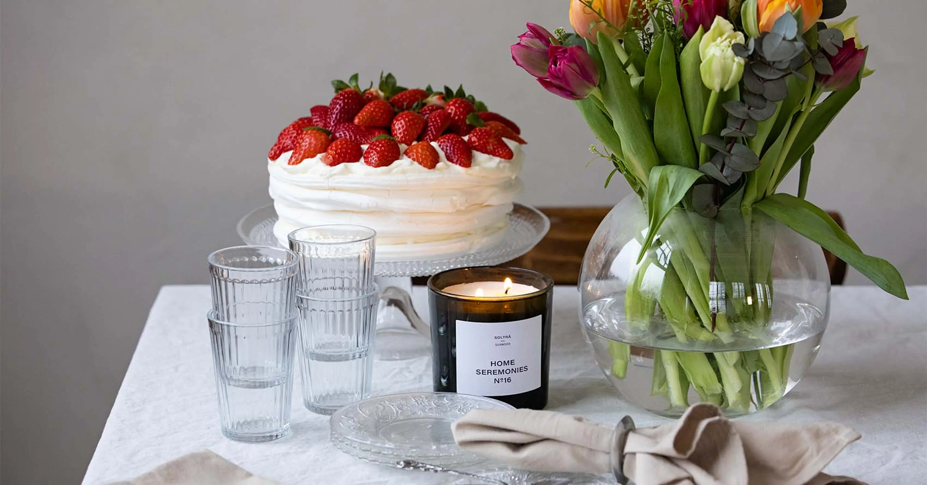 Ett svart doftljus märkt "Home Seremonies" brinner bredvid en rund glasvas med gröna stjälkar, omgivna av klara glas och en vacker glasskål på ett ljust dukat bord.