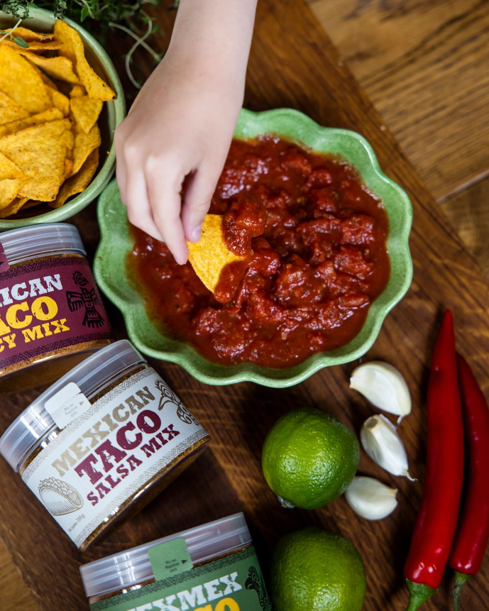En hand doppar en tortillachips i en grön skål med röd salsa. Runtom ligger kryddburkar, limefrukter, vitlök och röda chilipeppar.