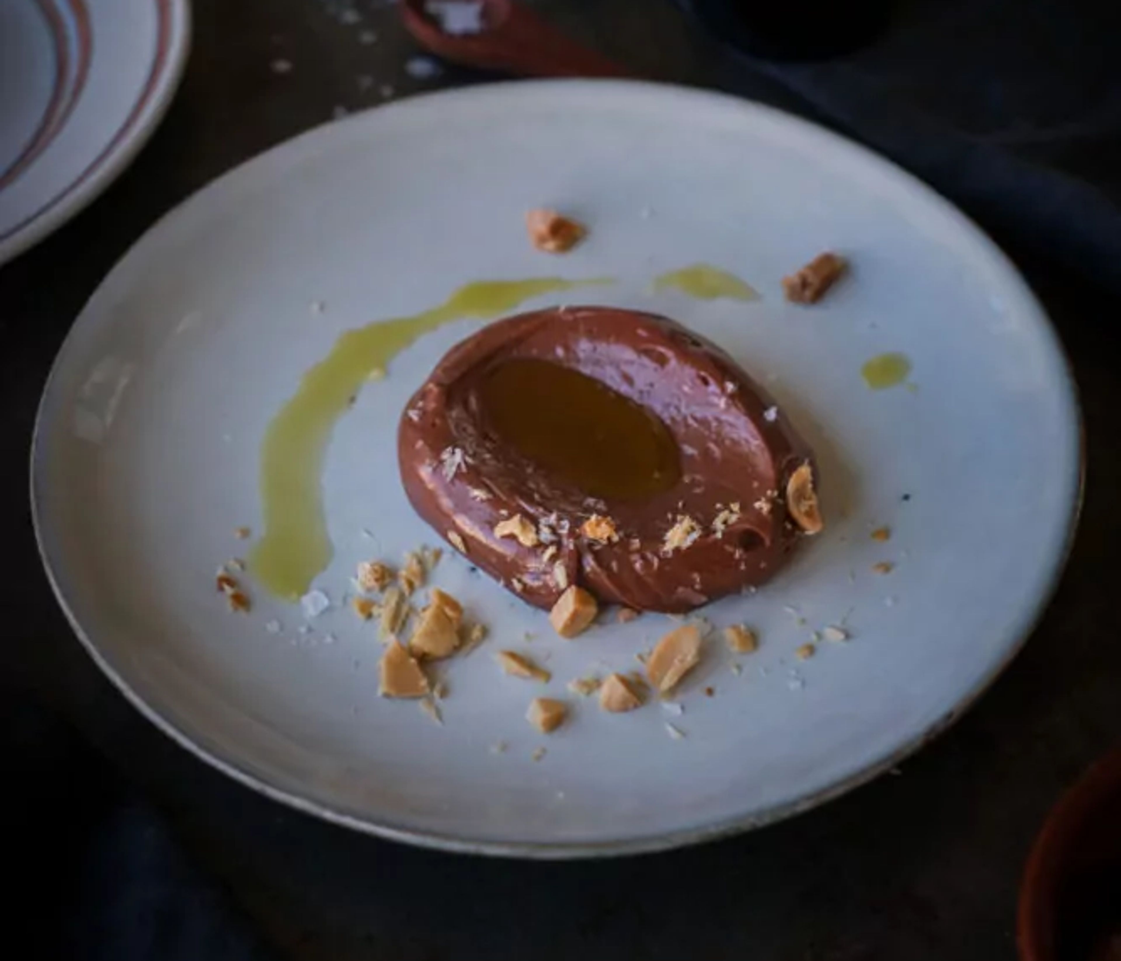 En klick len chokladkräm med en glansig olivolja i mitten, toppad med grovhackade nötbitar och ett stänk av flingsalt på en vit tallrik. I bakgrunden står en flaska extra virgin olivolja och en liten skål