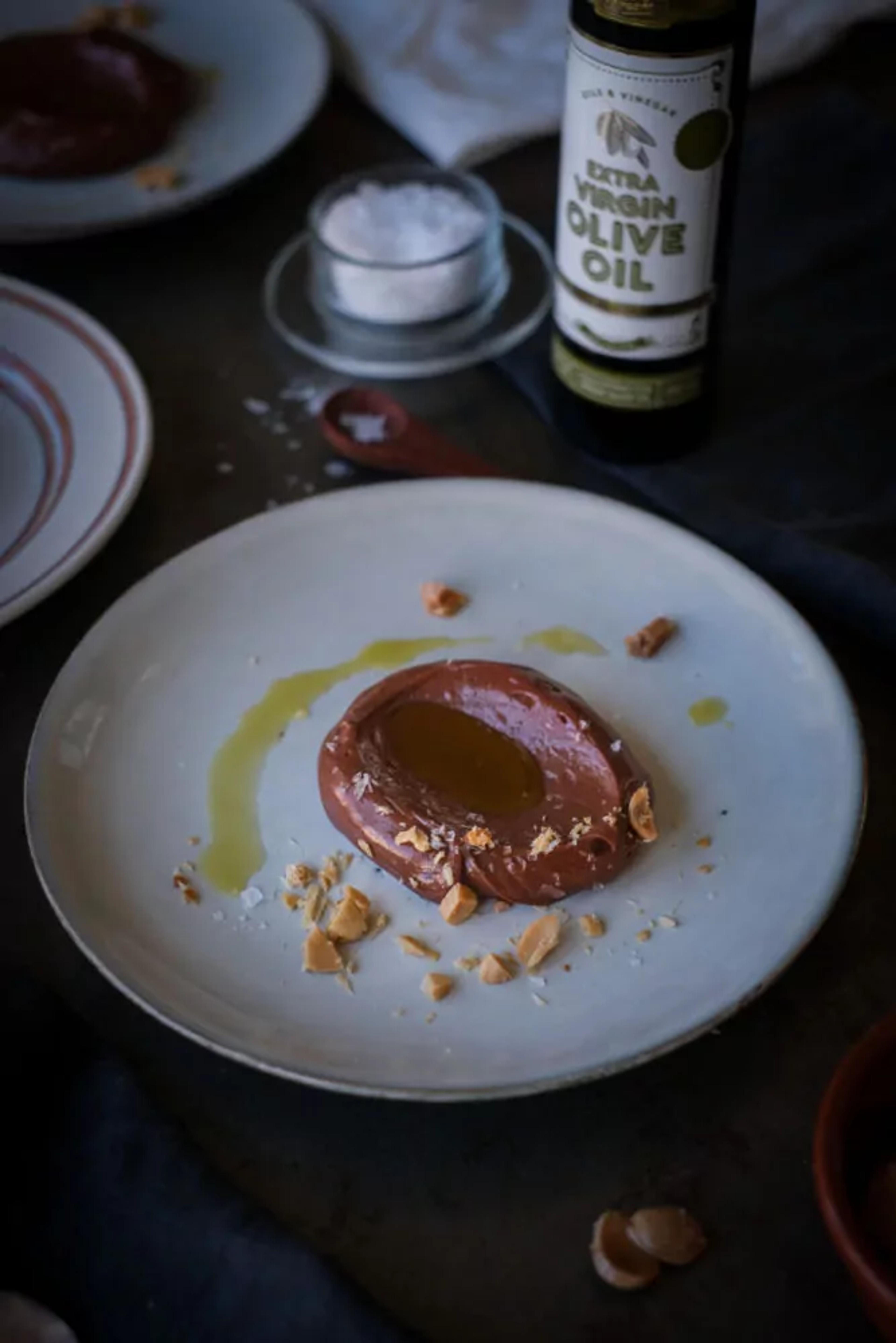 En klick len chokladkräm med en glansig olivolja i mitten, toppad med grovhackade nötbitar och ett stänk av flingsalt på en vit tallrik. I bakgrunden står en flaska extra virgin olivolja och en liten skål