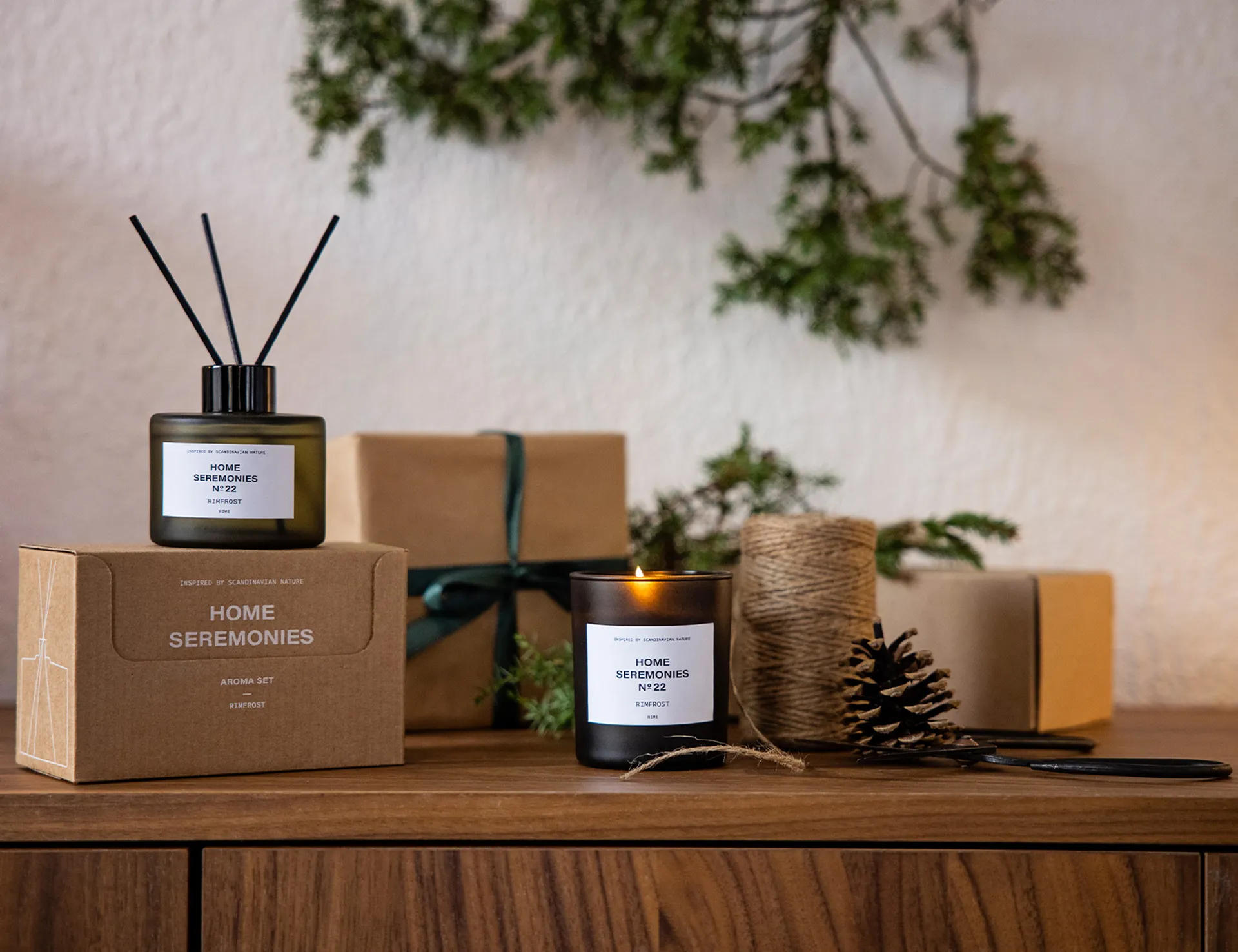 Ett doftset med rökelsepinnar i en grågrön glasflaska och ett tänd doftljus i matchande mörkt glas, båda märkta med "Rimfrost". Paket och julpynt som kottar, snöre och