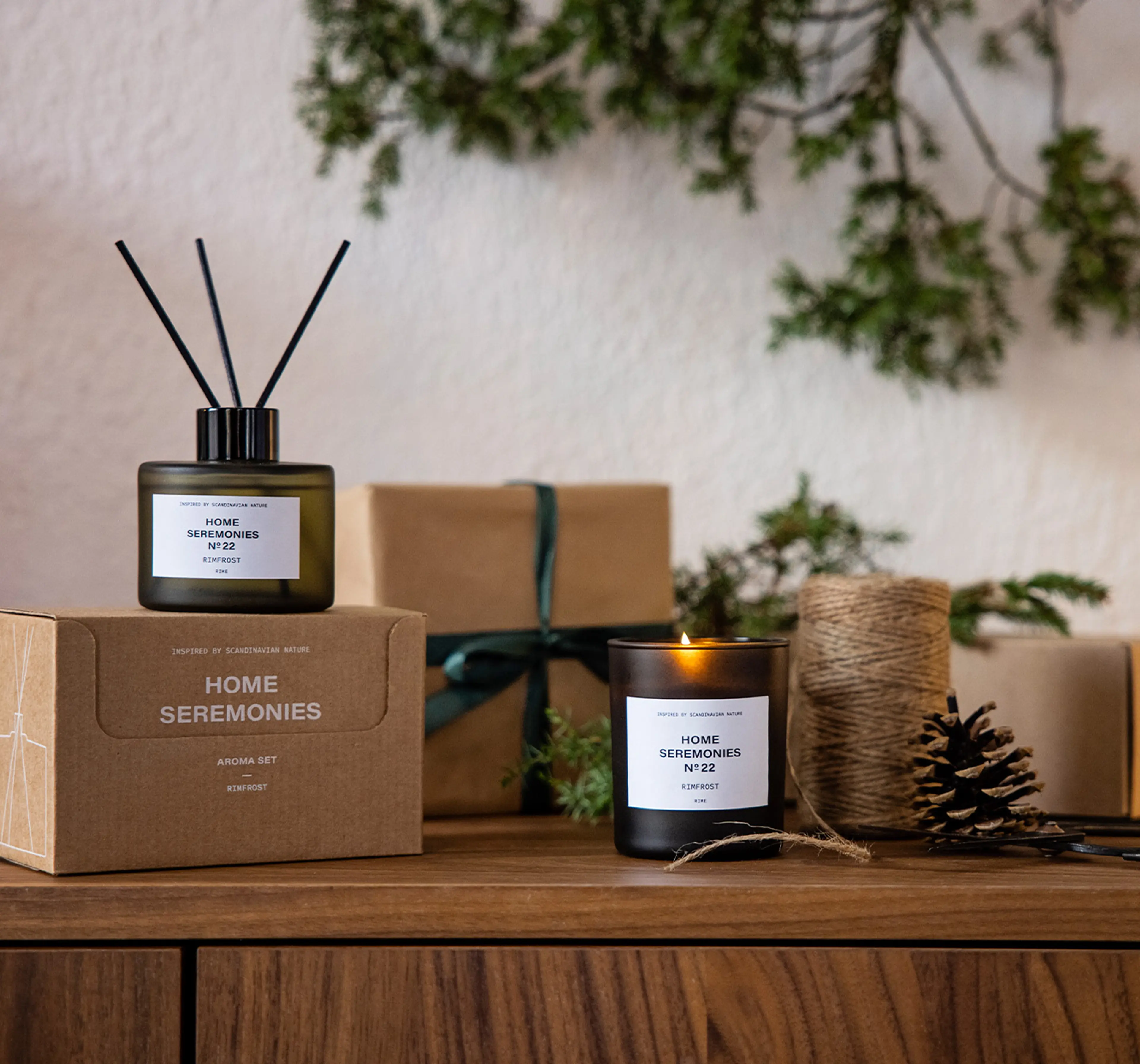 Ett doftset med rökelsepinnar i en grågrön glasflaska och ett tänd doftljus i matchande mörkt glas, båda märkta med "Rimfrost". Paket och julpynt som kottar, snöre och