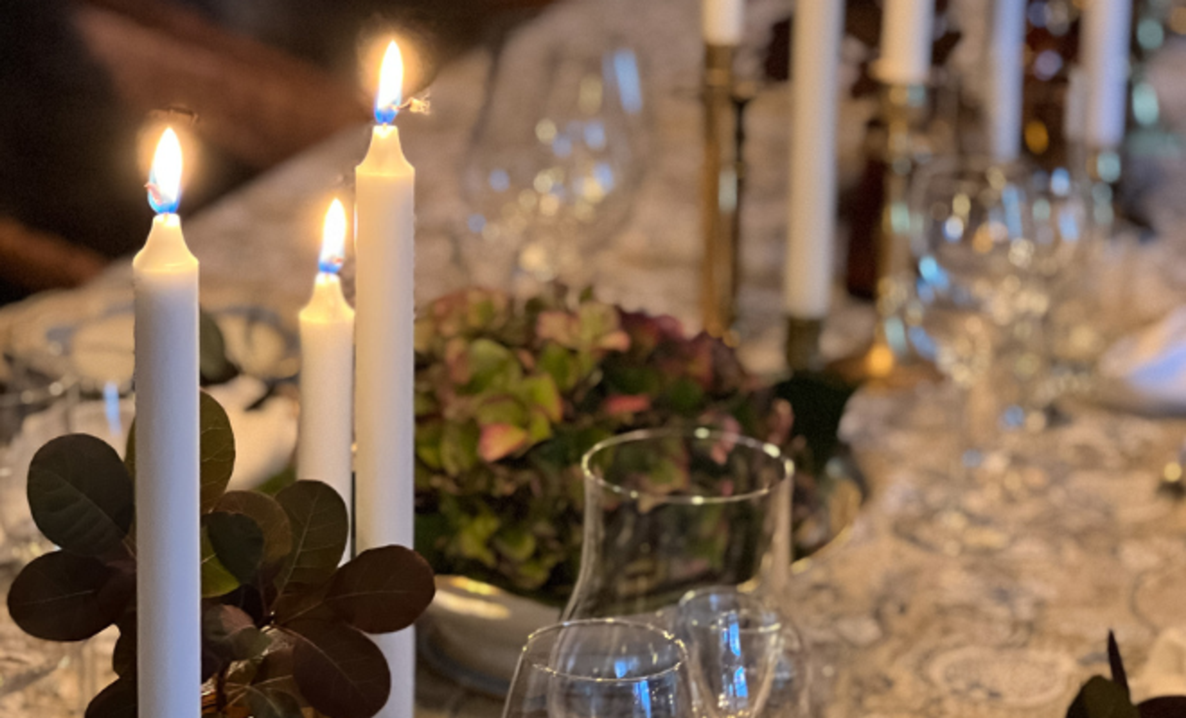 Tända vita ljus i eleganta ljusstakar på ett dukat bord med glas och gröna blad som dekoration. En inbjudande och detaljerad bordsmiljö för en festlig middag.