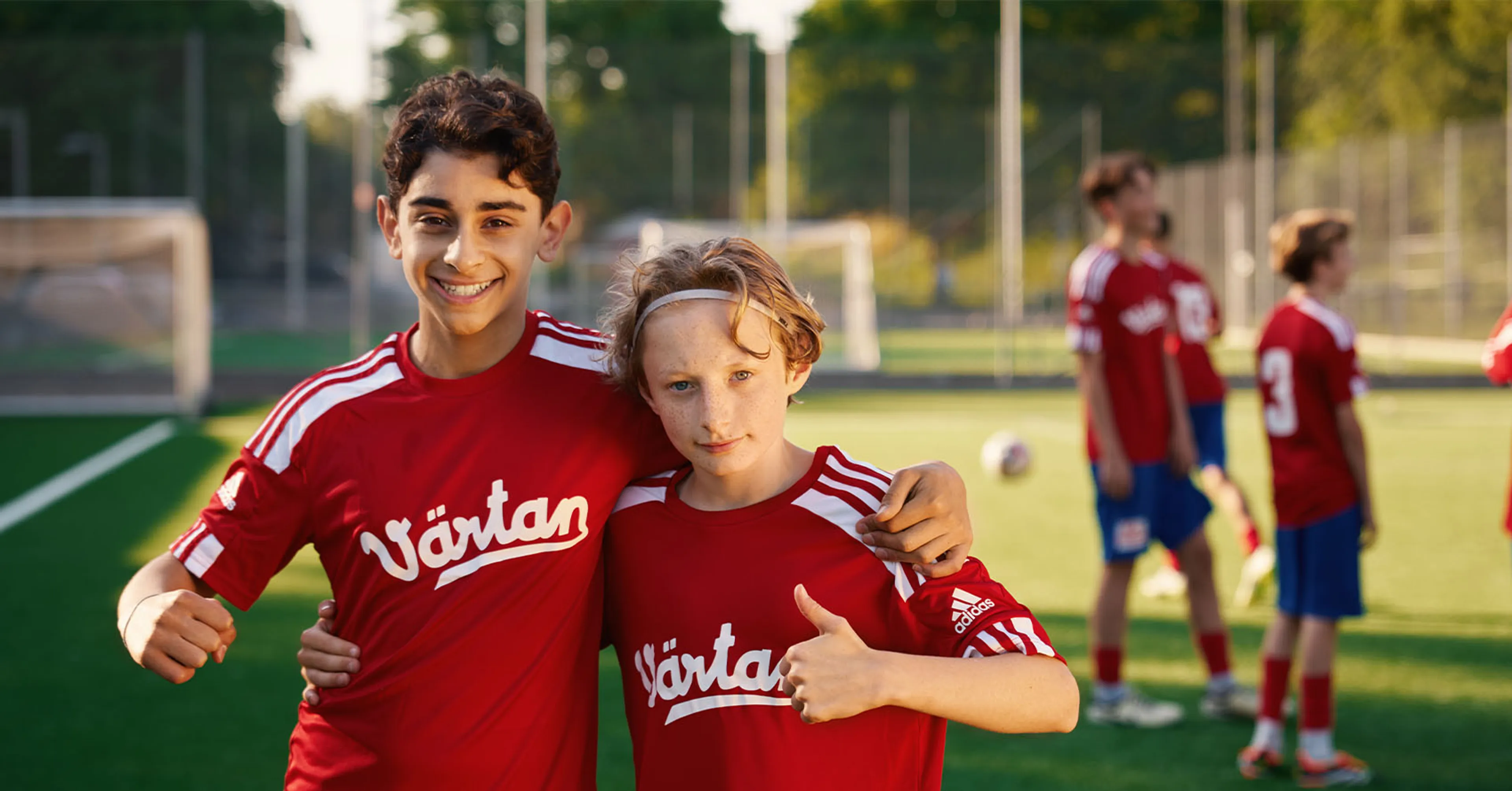 Två ungdomar i röda fotbollströjor med texten "Värtan" poserar tillsammans på en fotbollsplan i kvällsljus. I bakgrunden står fler lagkamrater i matchkläder.