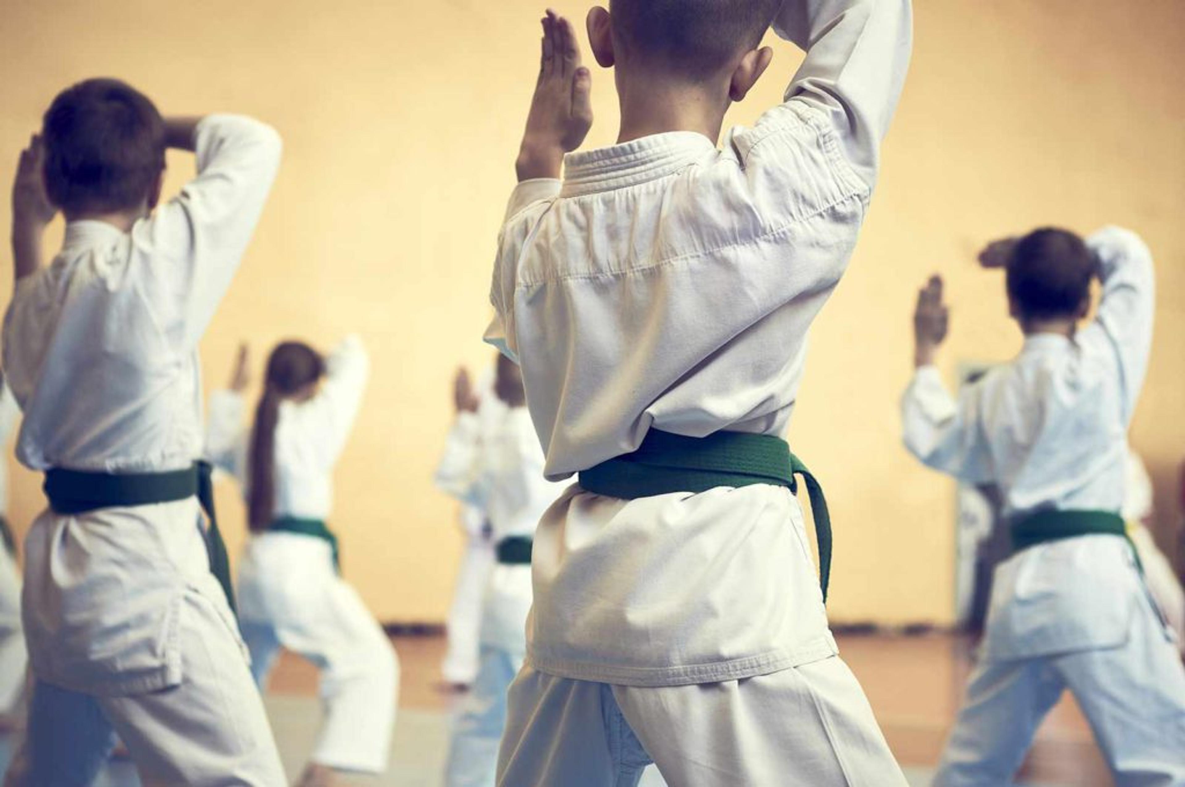 Barn i vita dräkter med gröna bälten tränar kampsport i en dojo. Gruppen fokuserar på en gemensam rörelse med händerna höjda.