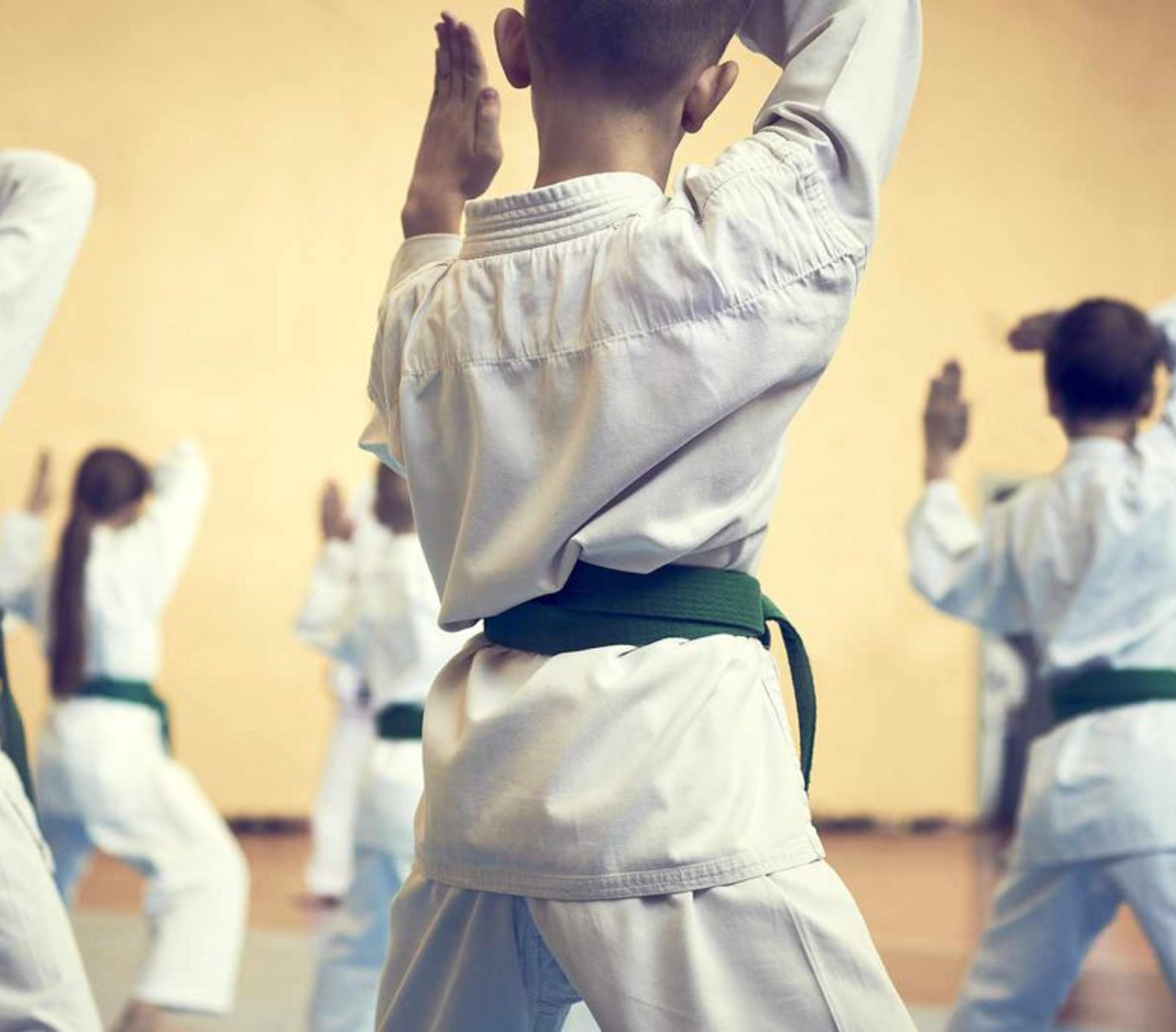 Barn i vita dräkter med gröna bälten tränar kampsport i en dojo. Gruppen fokuserar på en gemensam rörelse med händerna höjda.