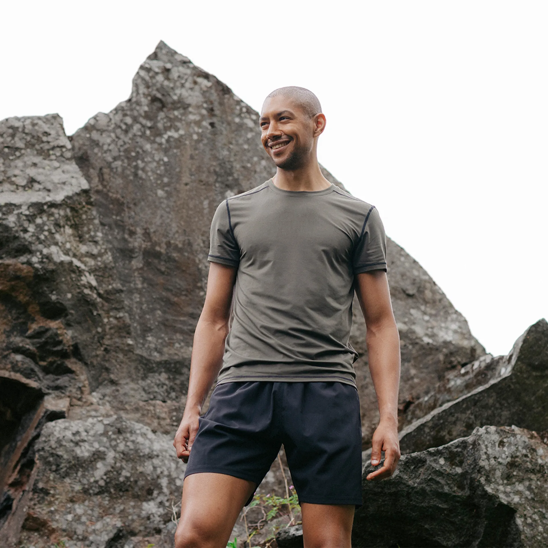 Mörkgrön funktions-t-shirt med kort ärm och kontrasterande sömmar, bärs av en leende person utomhus vid klippor. T-shirten har en enkel, lätt figursydd design för aktivt bruk.