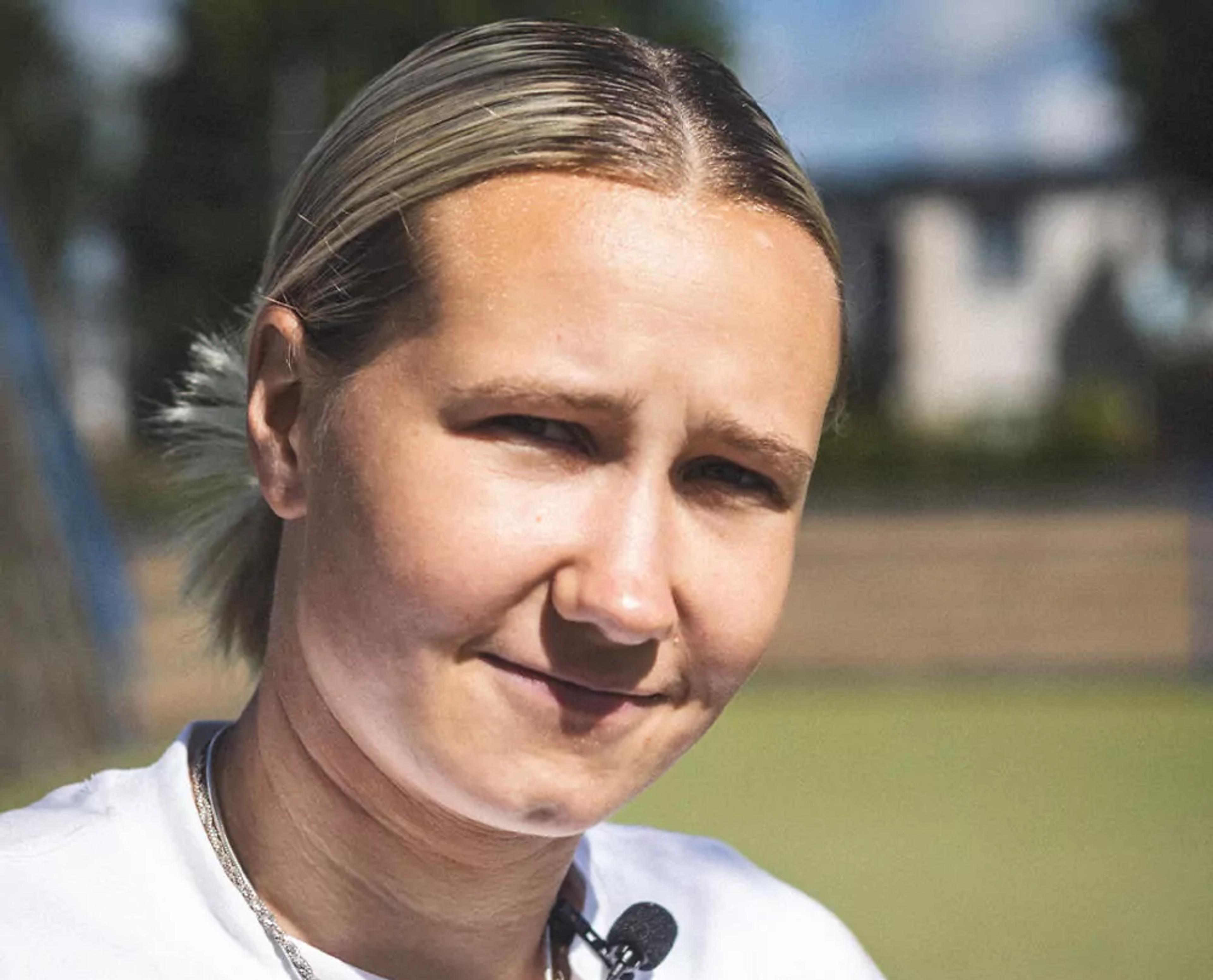 Person med ljust hår tillbakakammat i en tofs, iförd vit t-shirt och två tunna silverkedjor runt halsen. Personen bär en liten mikrofonfäste på bröstet.