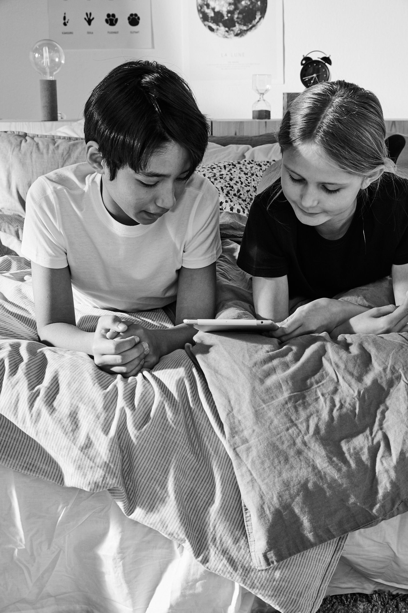 Två barn ligger tätt ihop i en säng och tittar på en surfplatta tillsammans. Rummet är personligt med tavlor och en väckarklocka i bakgrunden.