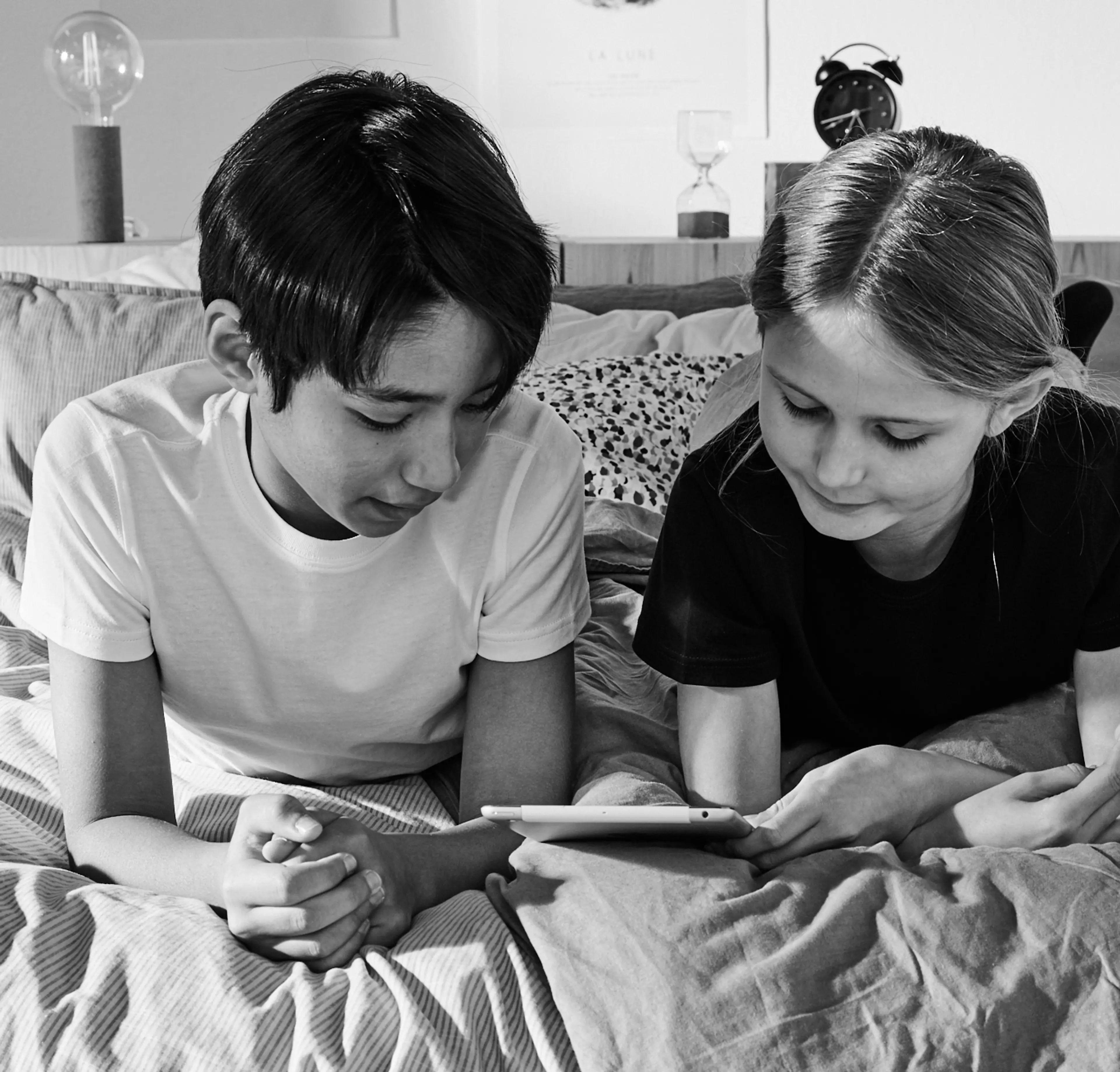 Två barn ligger tätt ihop i en säng och tittar på en surfplatta tillsammans. Rummet är personligt med tavlor och en väckarklocka i bakgrunden.