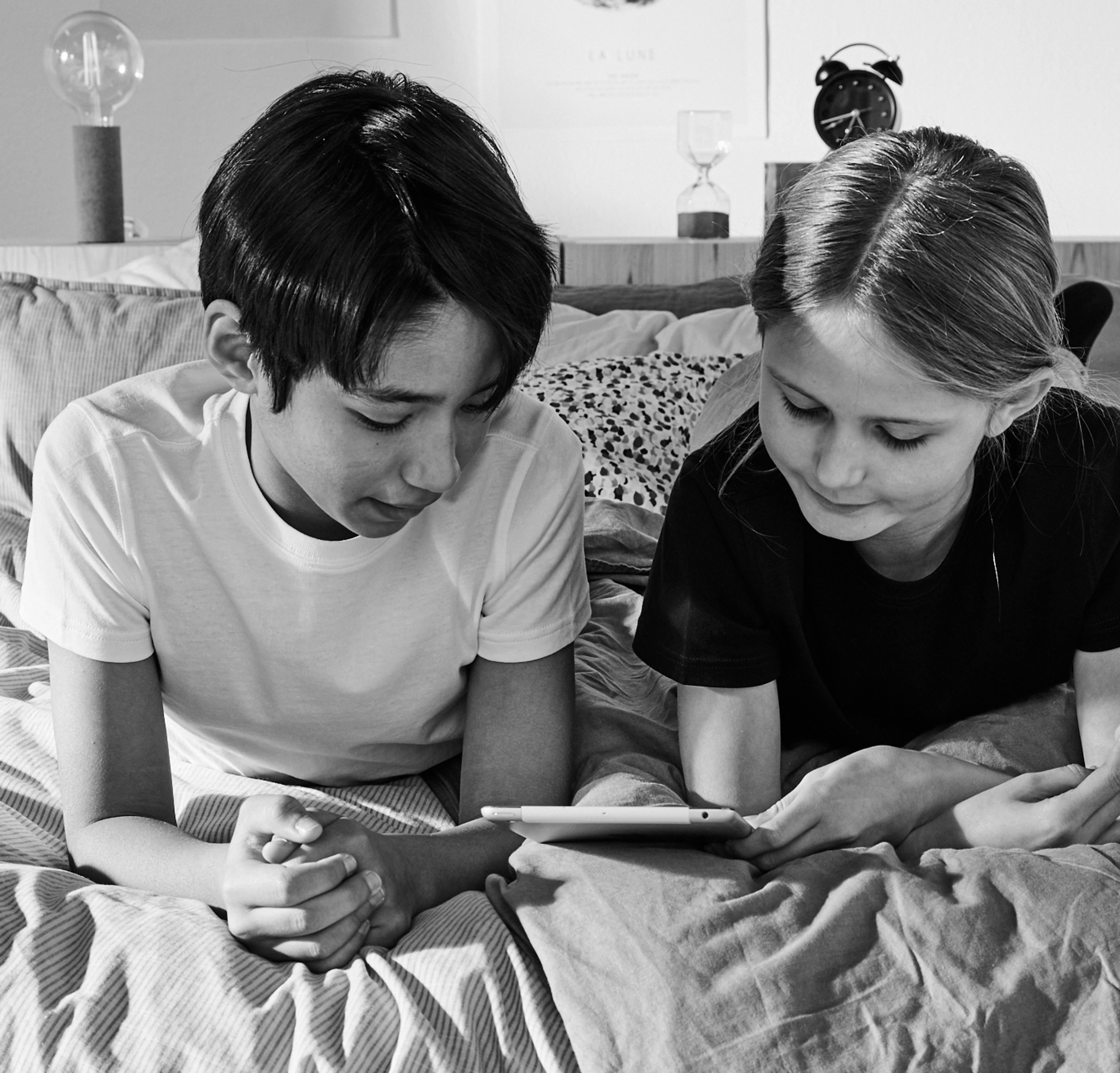 Två barn ligger tätt ihop i en säng och tittar på en surfplatta tillsammans. Rummet är personligt med tavlor och en väckarklocka i bakgrunden.