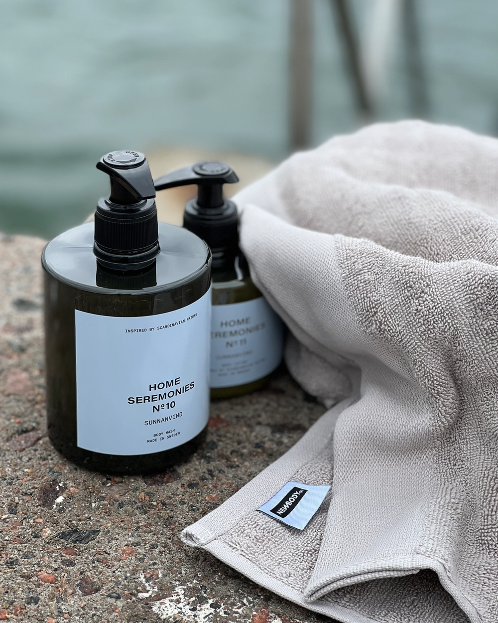 Två pumppåsflaskor med kroppstvål i mörkt plast, placerade på en stenbrygga vid vattnet tillsammans med en mjuk, ljusgrå handduk.