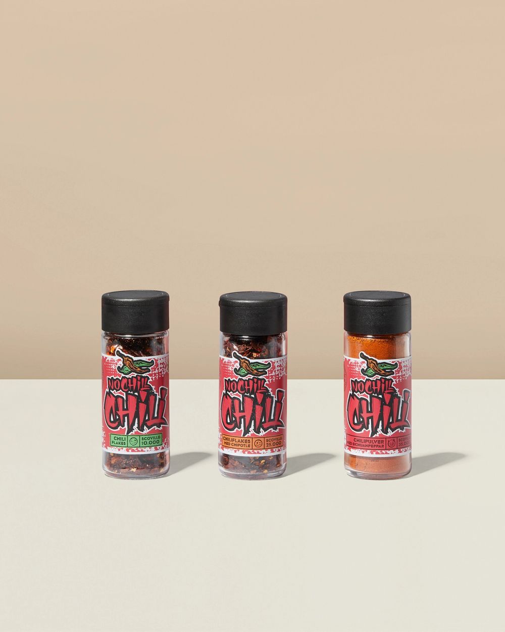 Tre glasburkar med chili i olika former – flakes och pulver – med svart lock och färgglada etiketter.