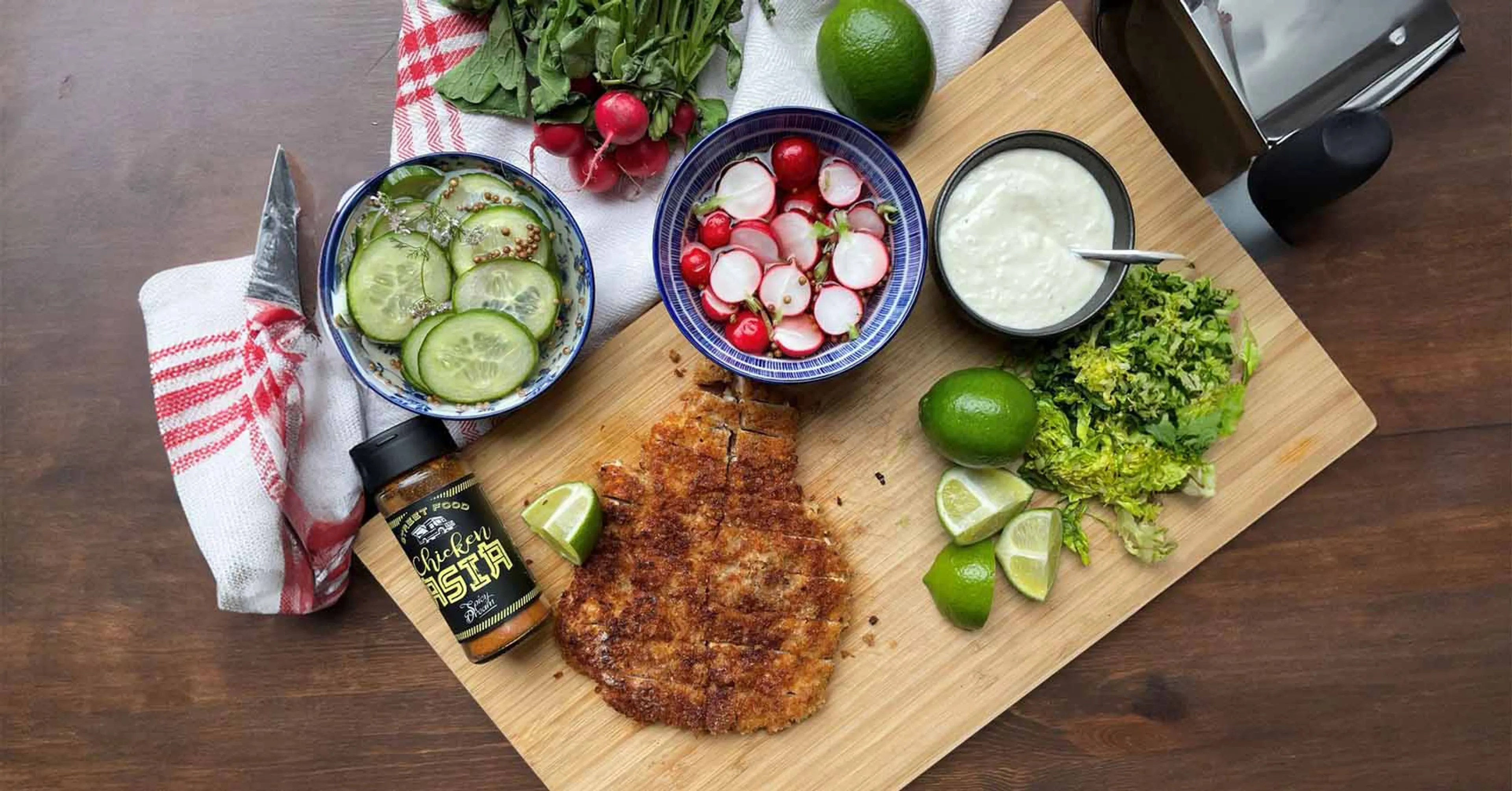 En krispig kycklingschnitzel med en limeklyfta på, serverad på ett skärbräde tillsammans med lime, en skål med sås och skivade grönsaker. På bordet finns också en portion ris och kryddburkar.