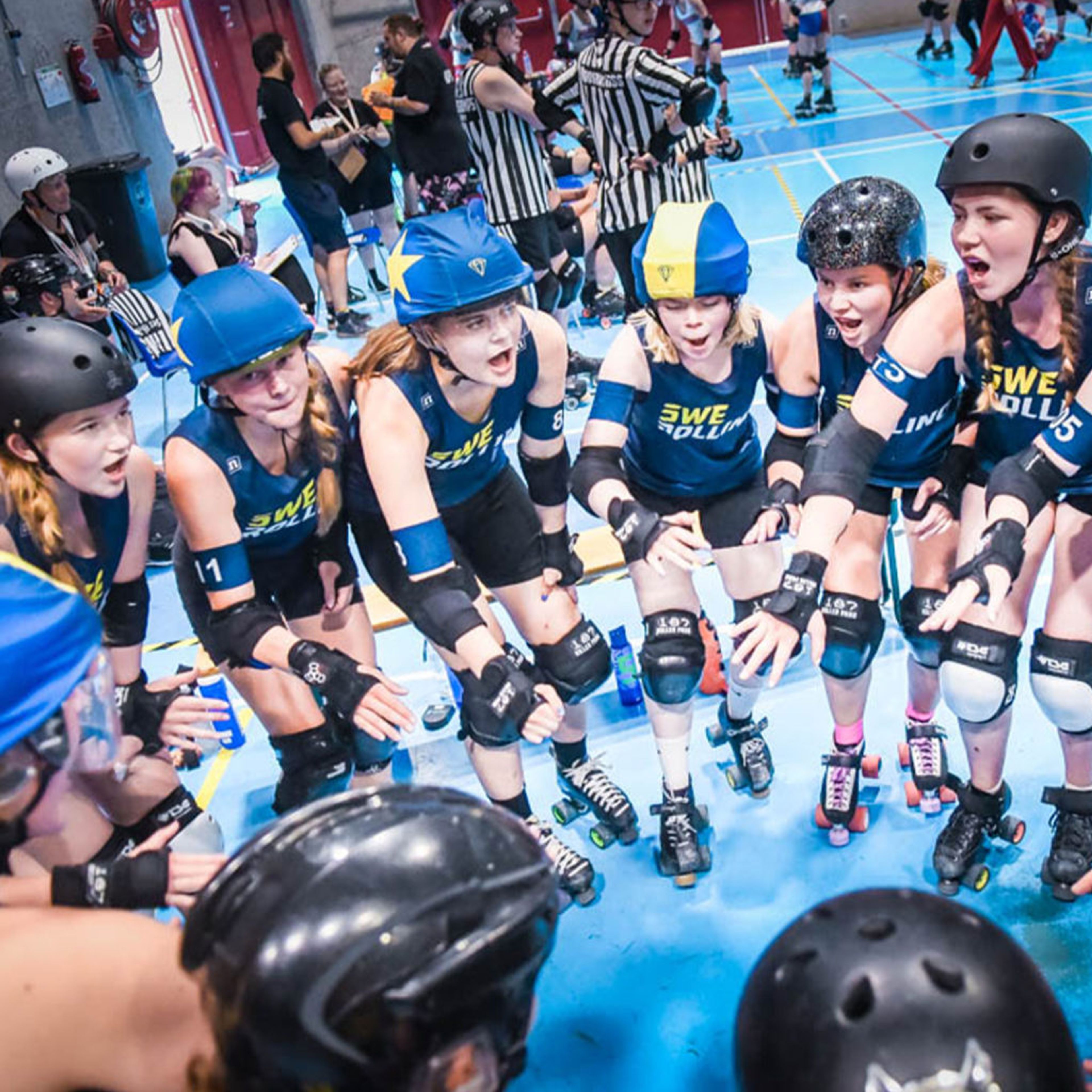 Juniorer i rullskridskor med blå och gula hjälmar samlade i en cirkel inför en match i roller derby. De bär skyddsutrustning och matchtröjor med texten "SWE".