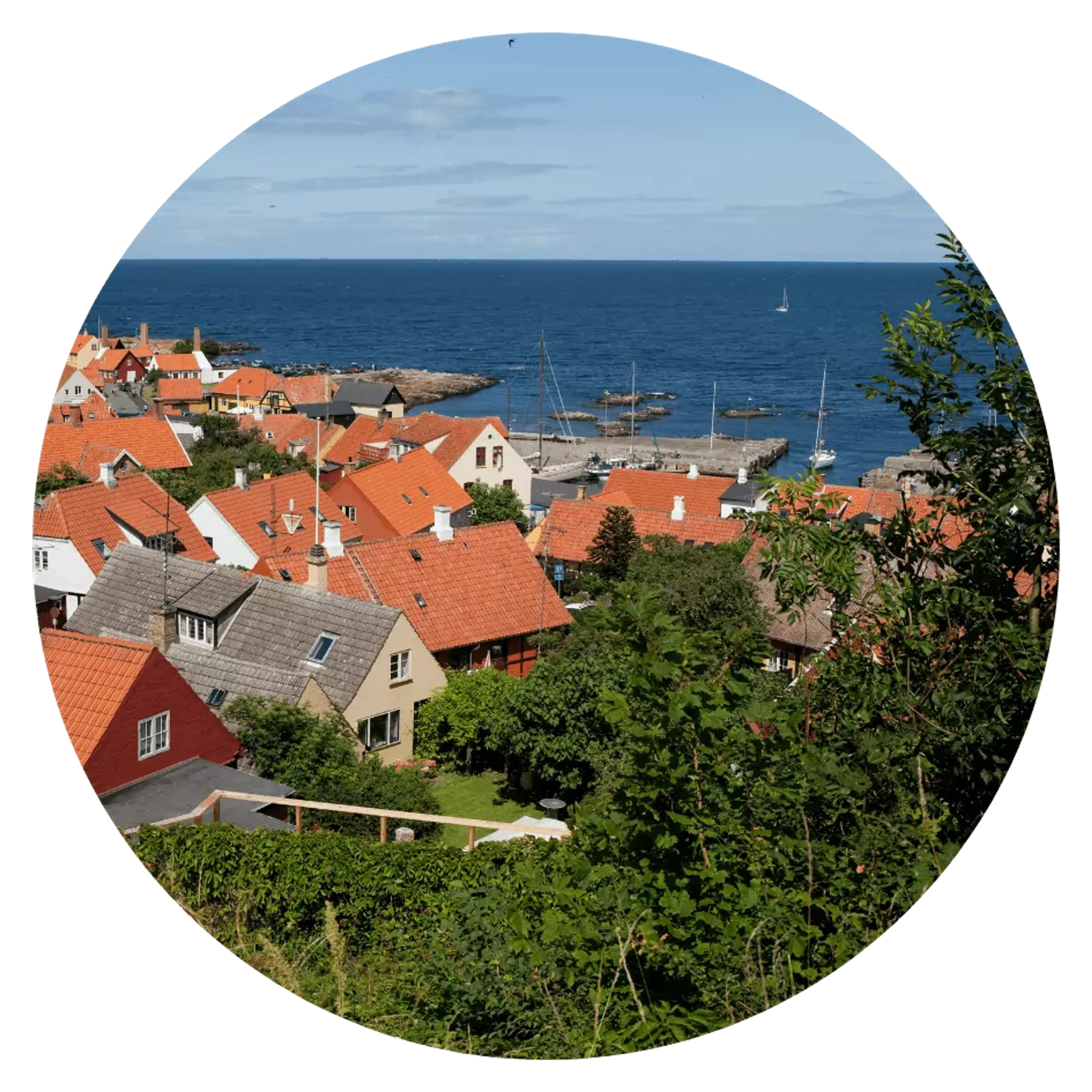 Ett kustsamhälle med röda tegeltak och små segelbåtar i hamnen bortom husen. Grönska och blått hav omger den pittoreska byn.