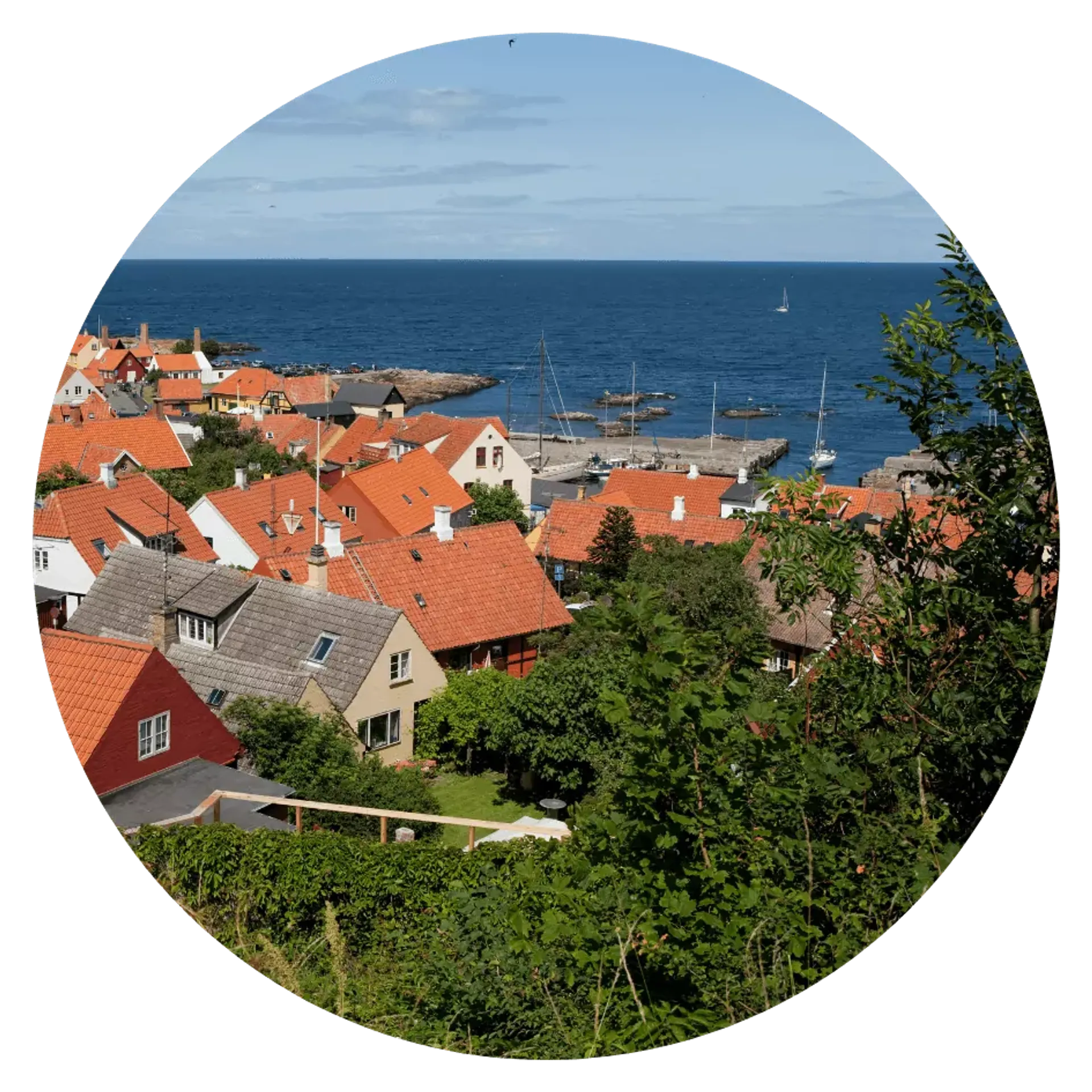 Ett kustsamhälle med röda tegeltak och små segelbåtar i hamnen bortom husen. Grönska och blått hav omger den pittoreska byn.