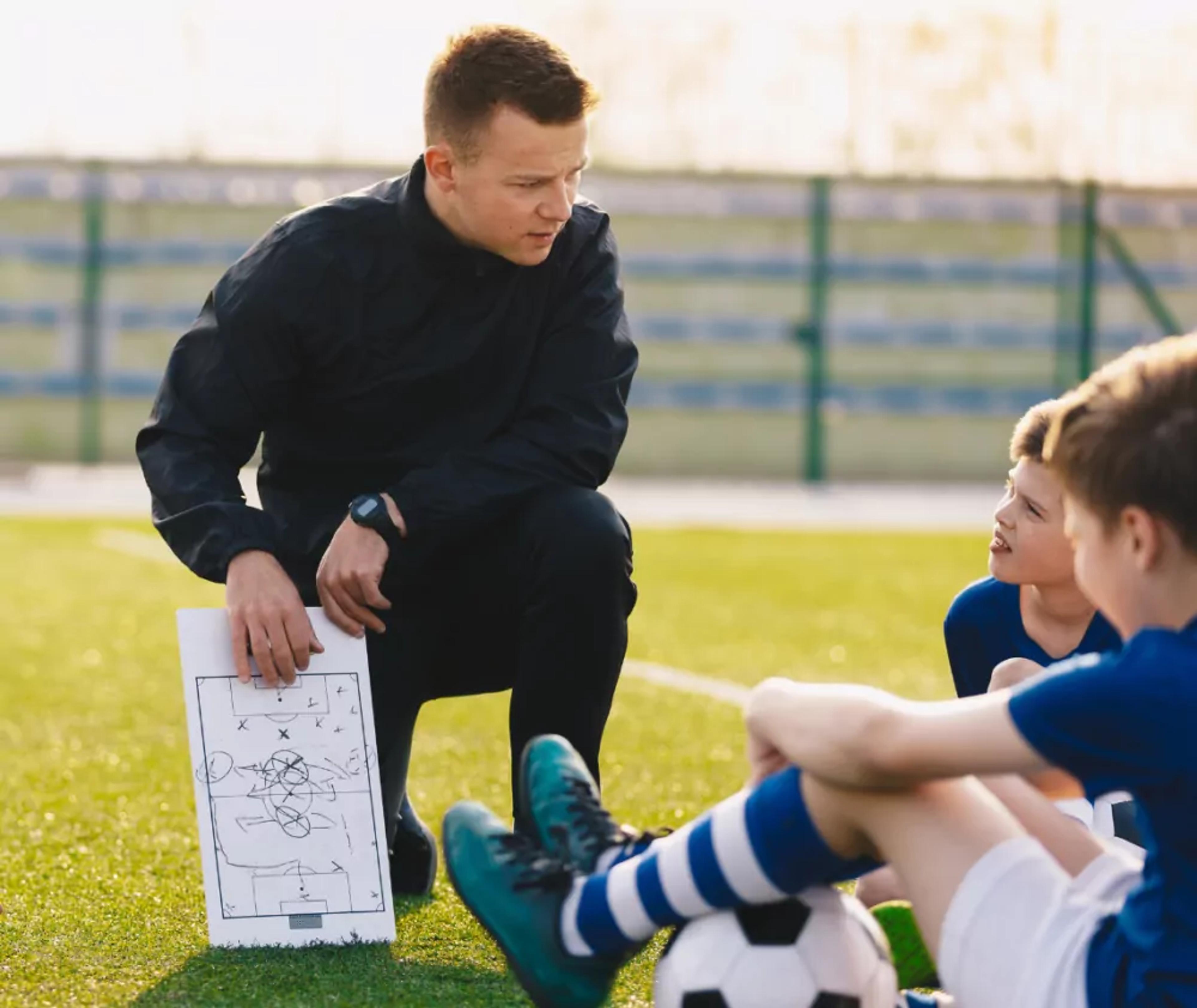 En ledare knäböjer på fotbollsplanen med en taktiktavla i handen och samtalar med två unga spelare i blå tröjor och randiga strumpor.