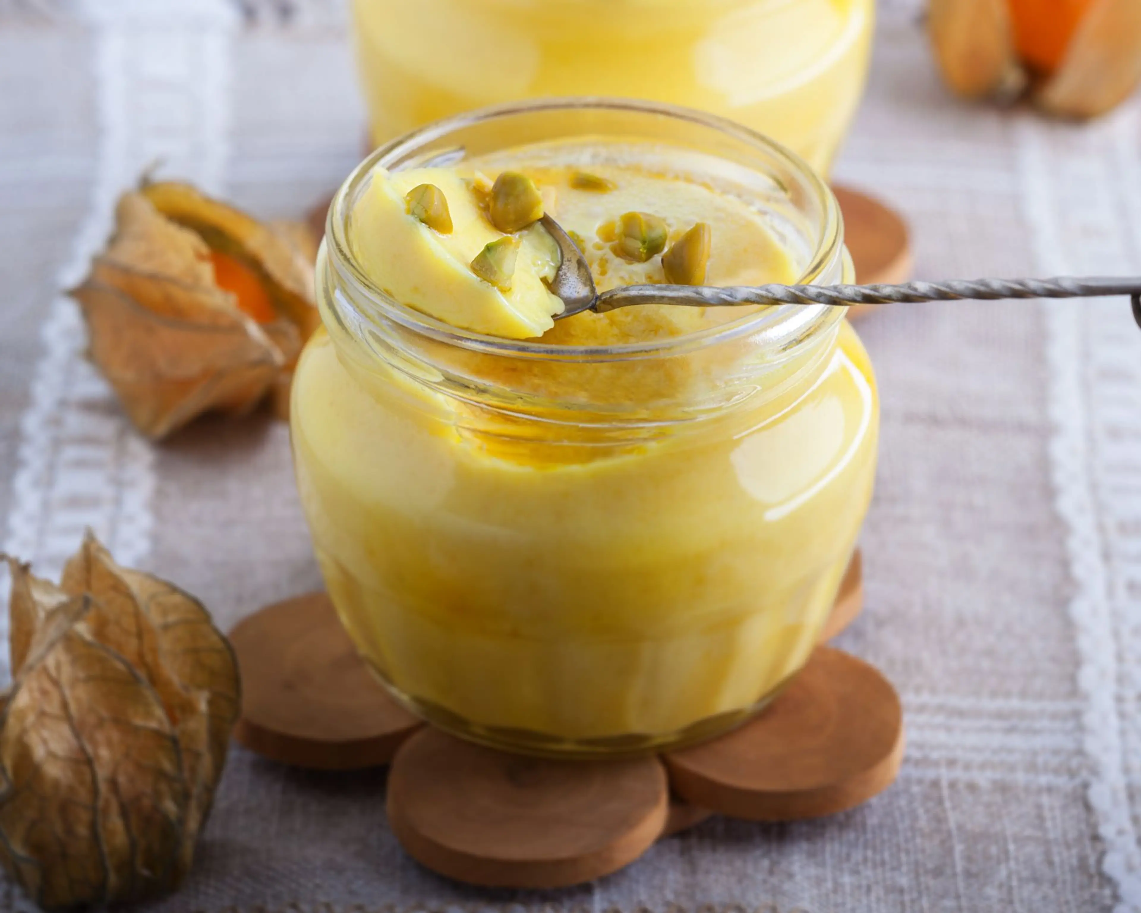 Krämig saffranspannacotta i en liten glasburk, garnerad med hackade pistagenötter. Burken står på ett träunderlägg på en linneduk med dekorativa physalis runtom.