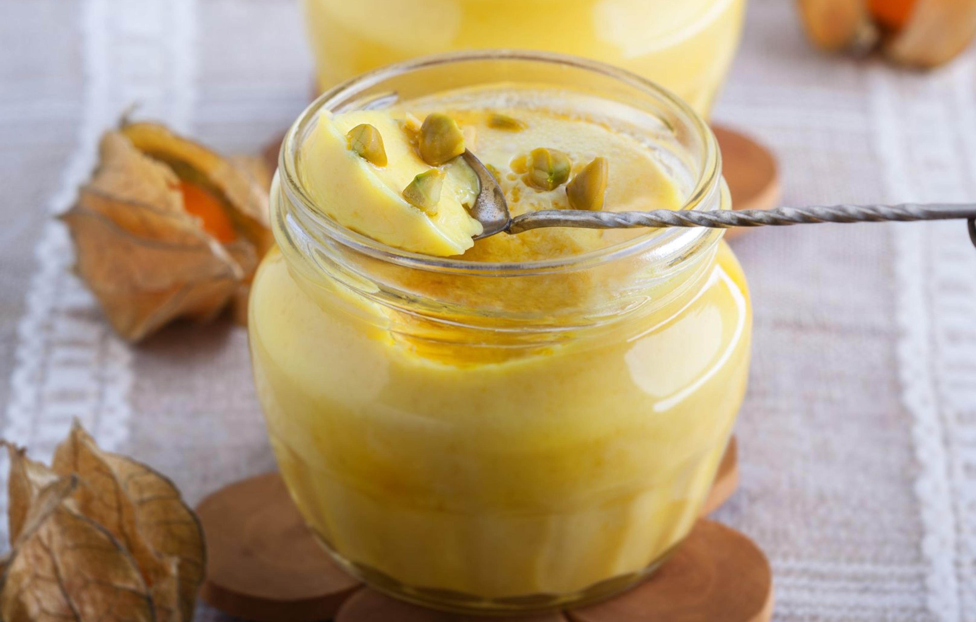 Krämig saffranspannacotta i en liten glasburk, garnerad med hackade pistagenötter. Burken står på ett träunderlägg på en linneduk med dekorativa physalis runtom.