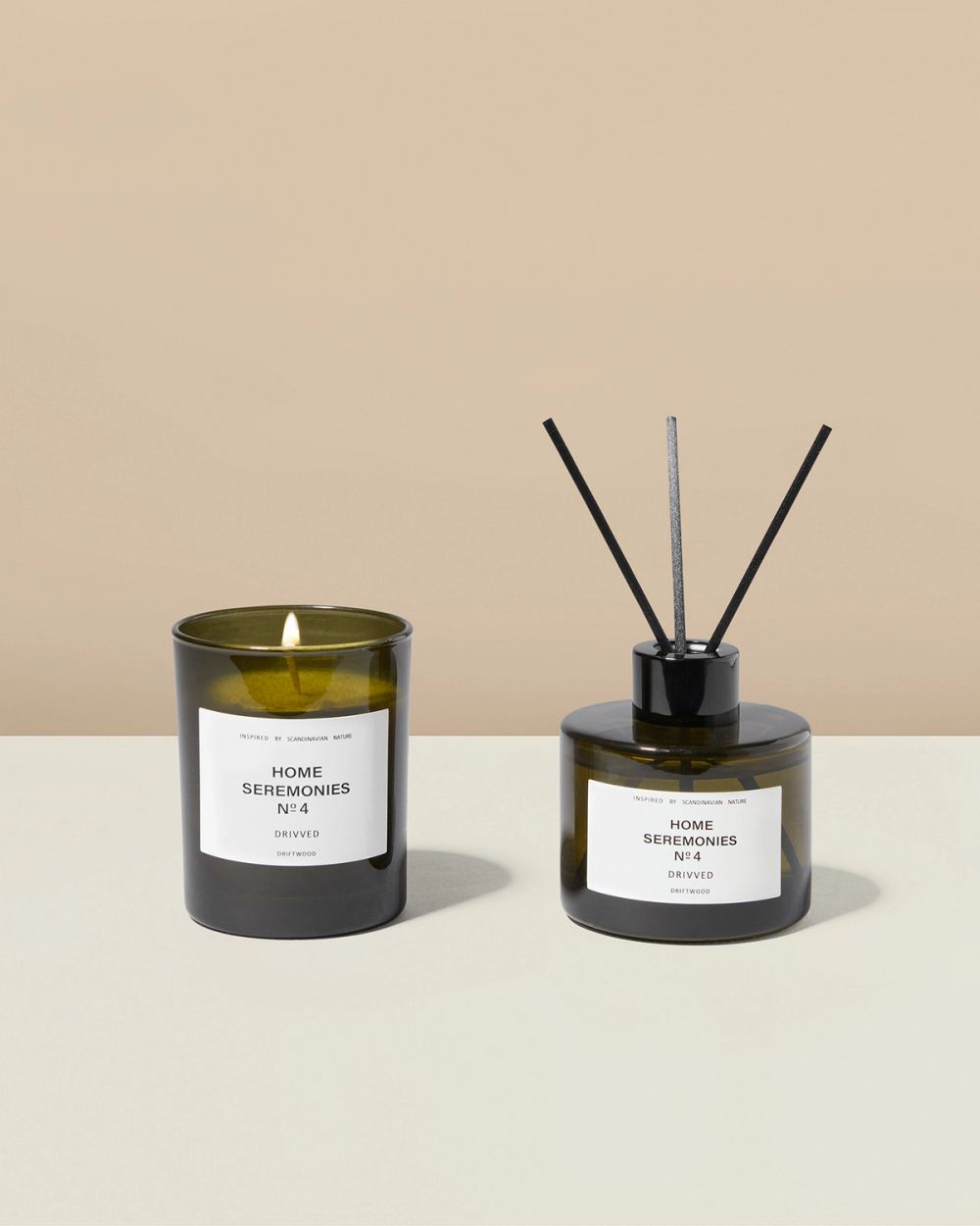 Ett doftljus med brinnande veke och en doftpinnarflaska i mörkgrönt glas med etikett i beige och svart text. Båda produkterna har doften drivved och är enkla i design.