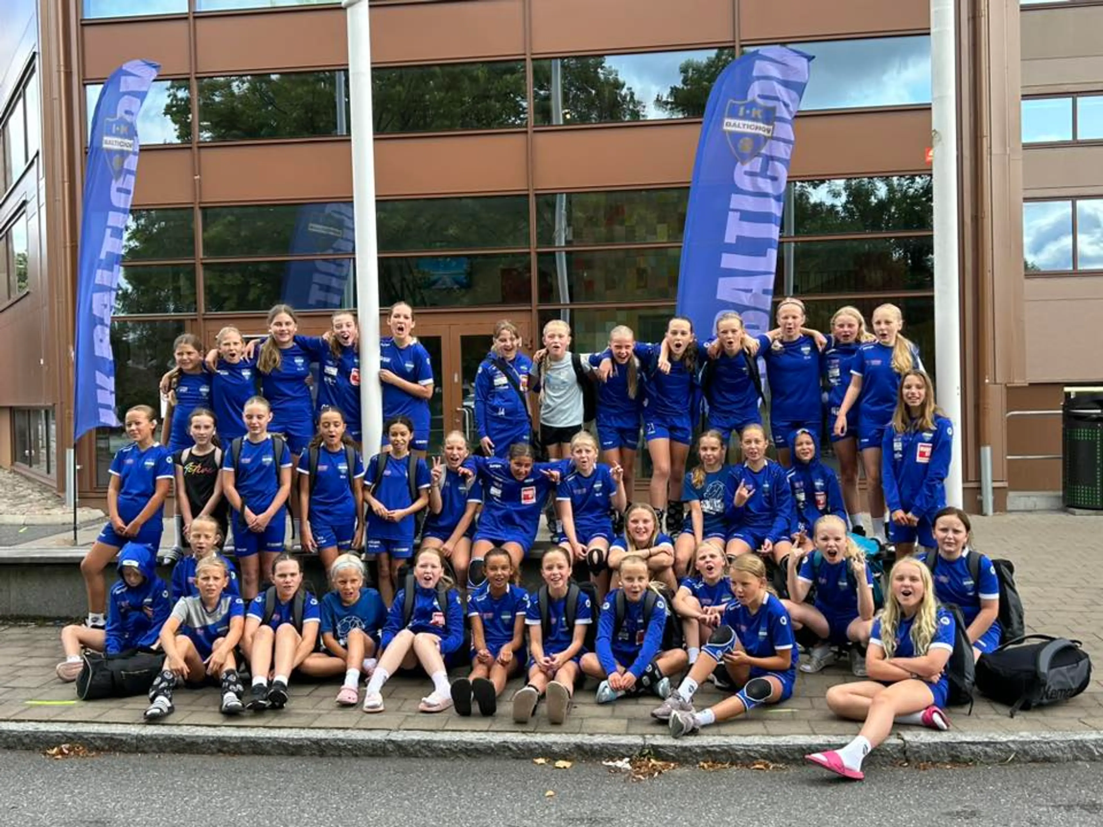 En stor grupp barn och ungdomar i blå sportkläder samlade utanför en byggnad med två blåa vimplar med texten "IK Baltichov". Gruppen verkar redo för laget eller en aktivitet tillsammans.
