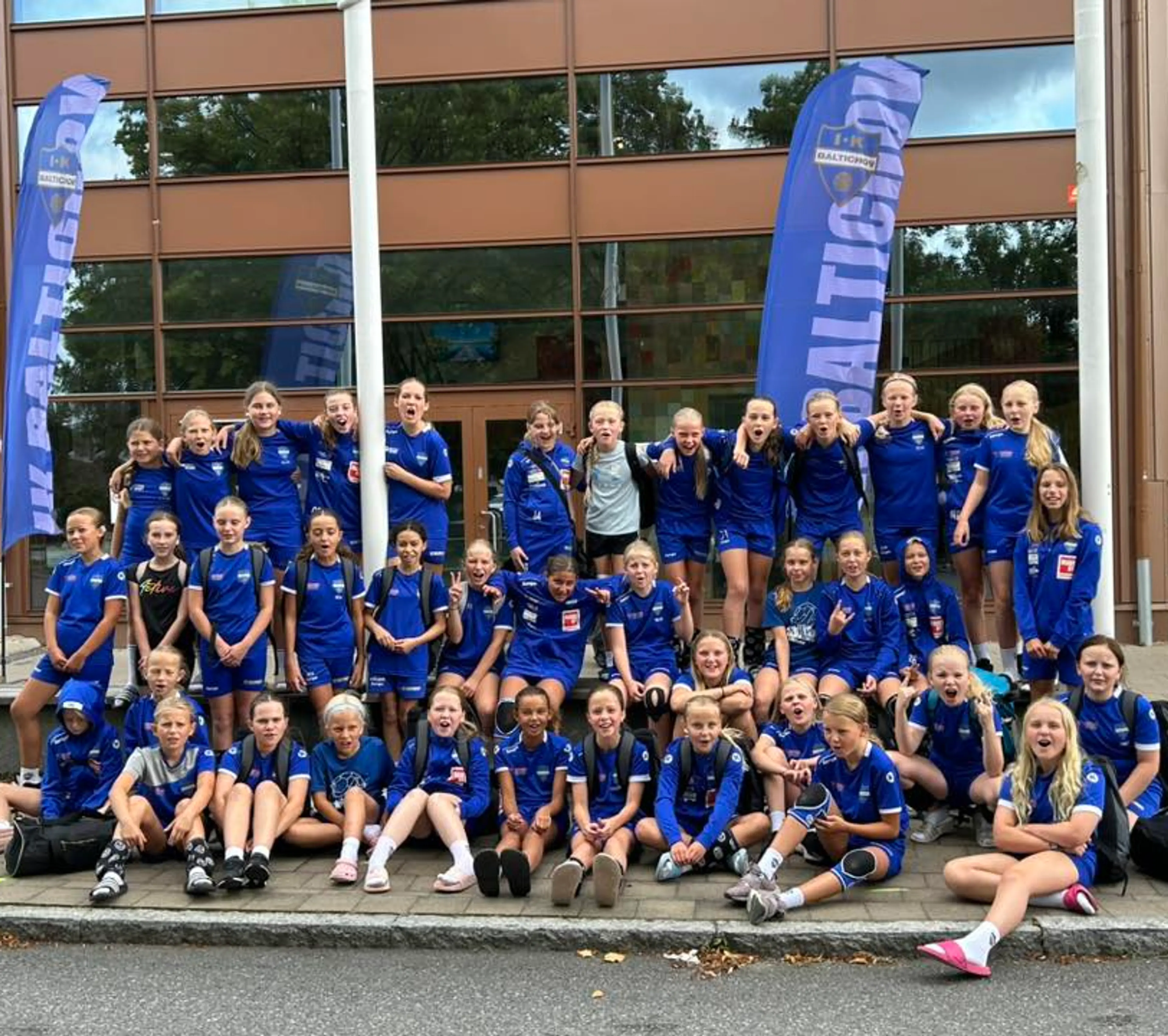 En stor grupp barn och ungdomar i blå sportkläder samlade utanför en byggnad med två blåa vimplar med texten "IK Baltichov". Gruppen verkar redo för laget eller en aktivitet tillsammans.