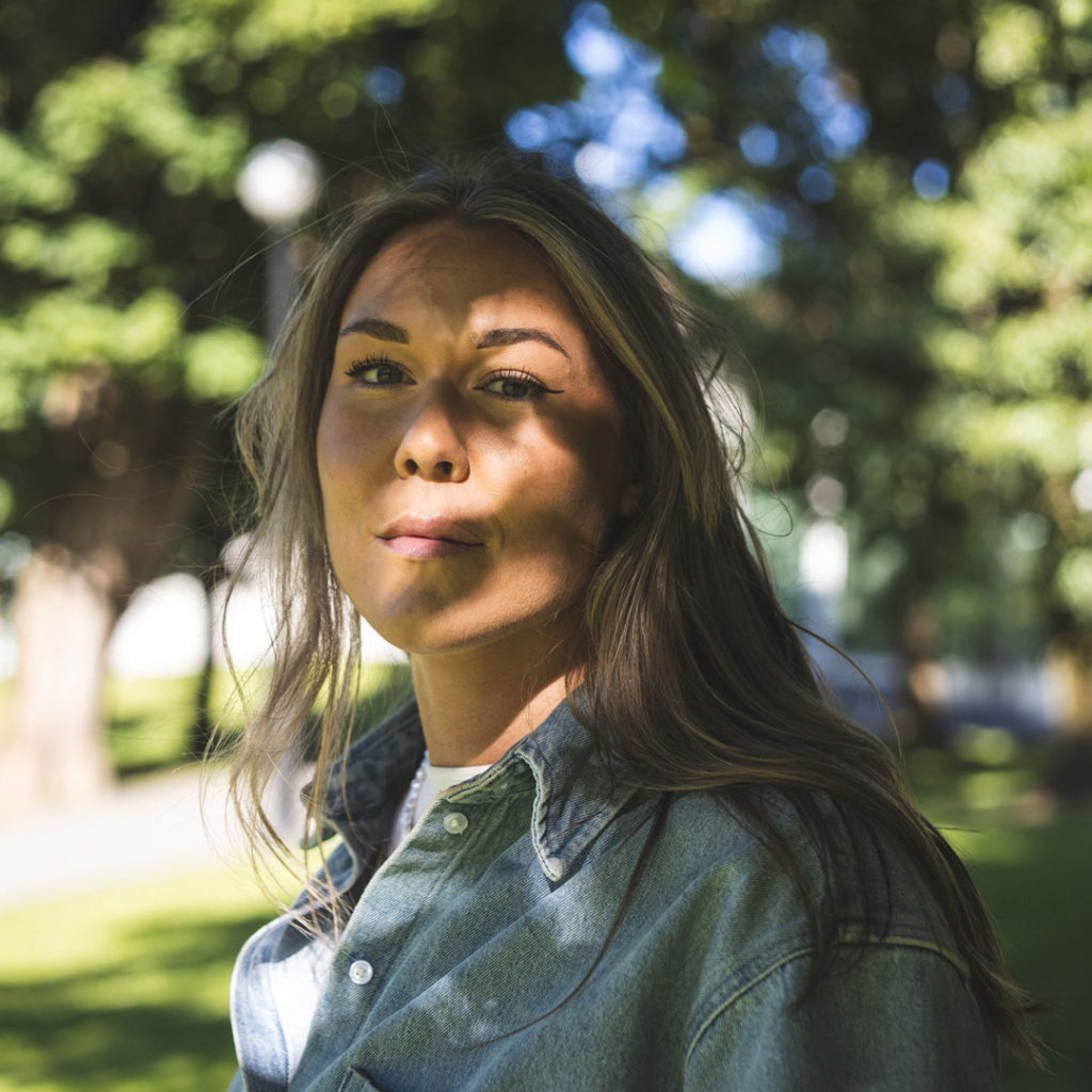 En person med långt hår och jeansskjorta står utomhus med gröna träd i bakgrunden. Solens strålar skapar skuggmönster över hennes ansikte.