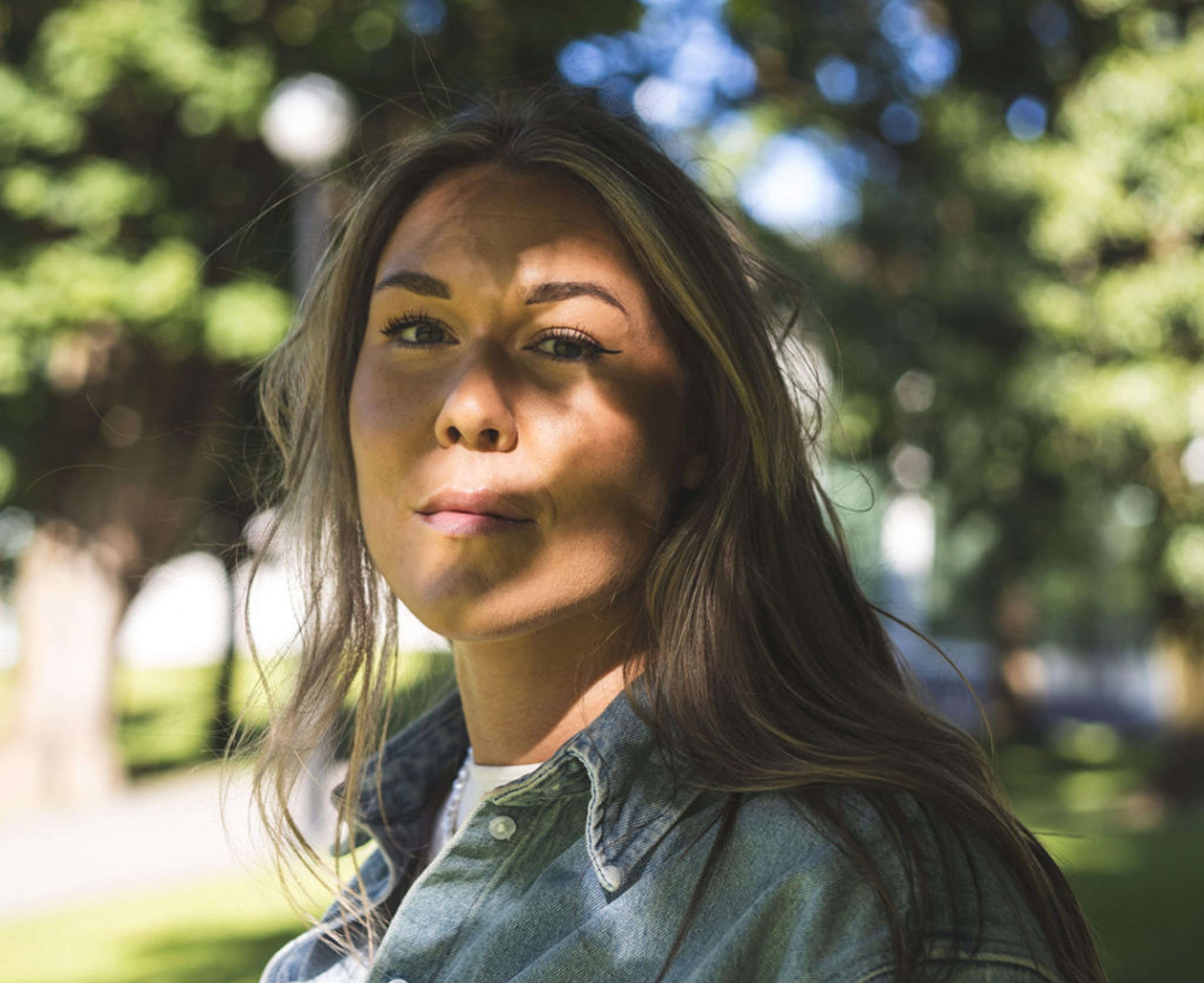 En person med långt hår och jeansskjorta står utomhus med gröna träd i bakgrunden. Solens strålar skapar skuggmönster över hennes ansikte.