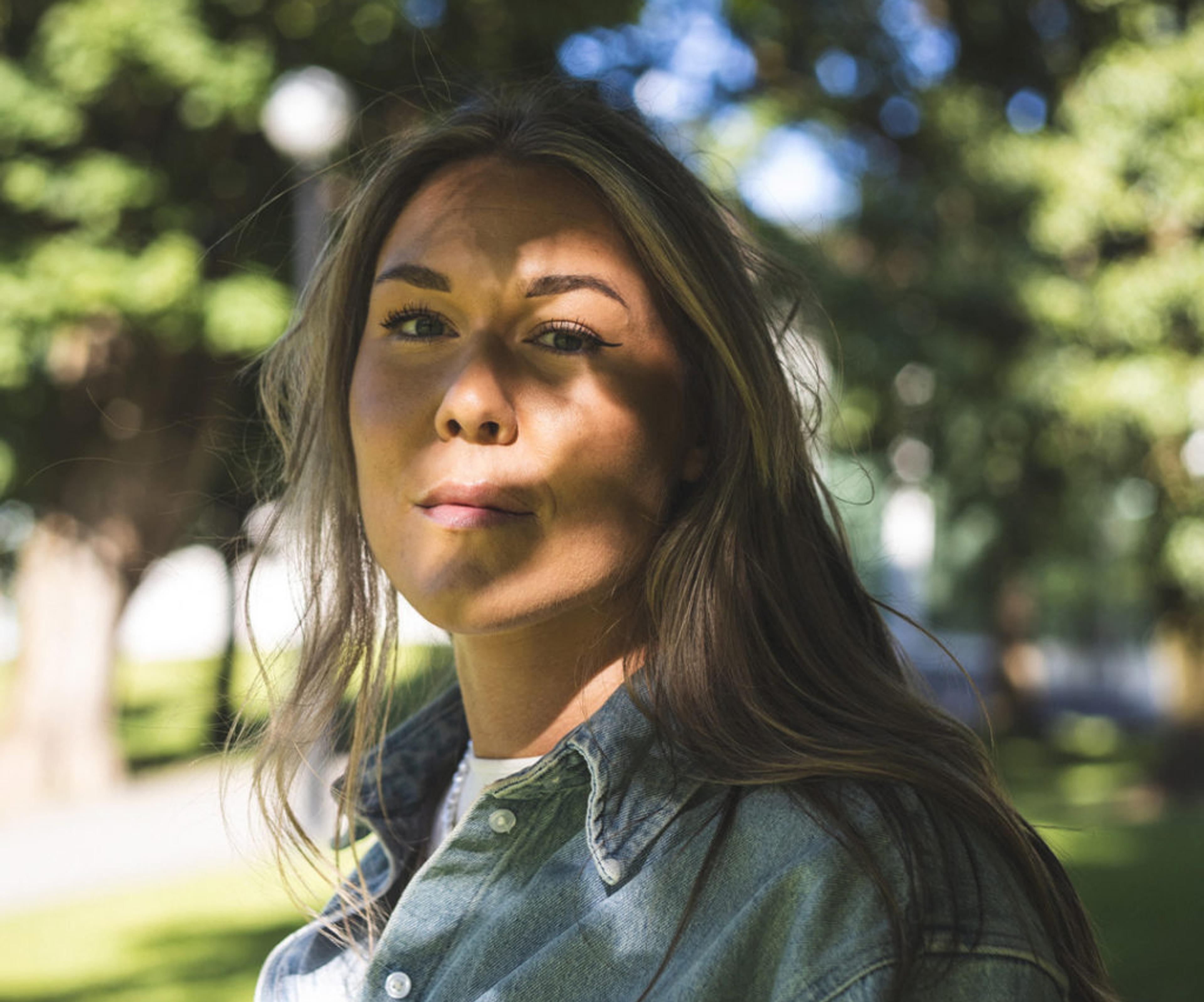 En person med långt hår och jeansskjorta står utomhus med gröna träd i bakgrunden. Solens strålar skapar skuggmönster över hennes ansikte.