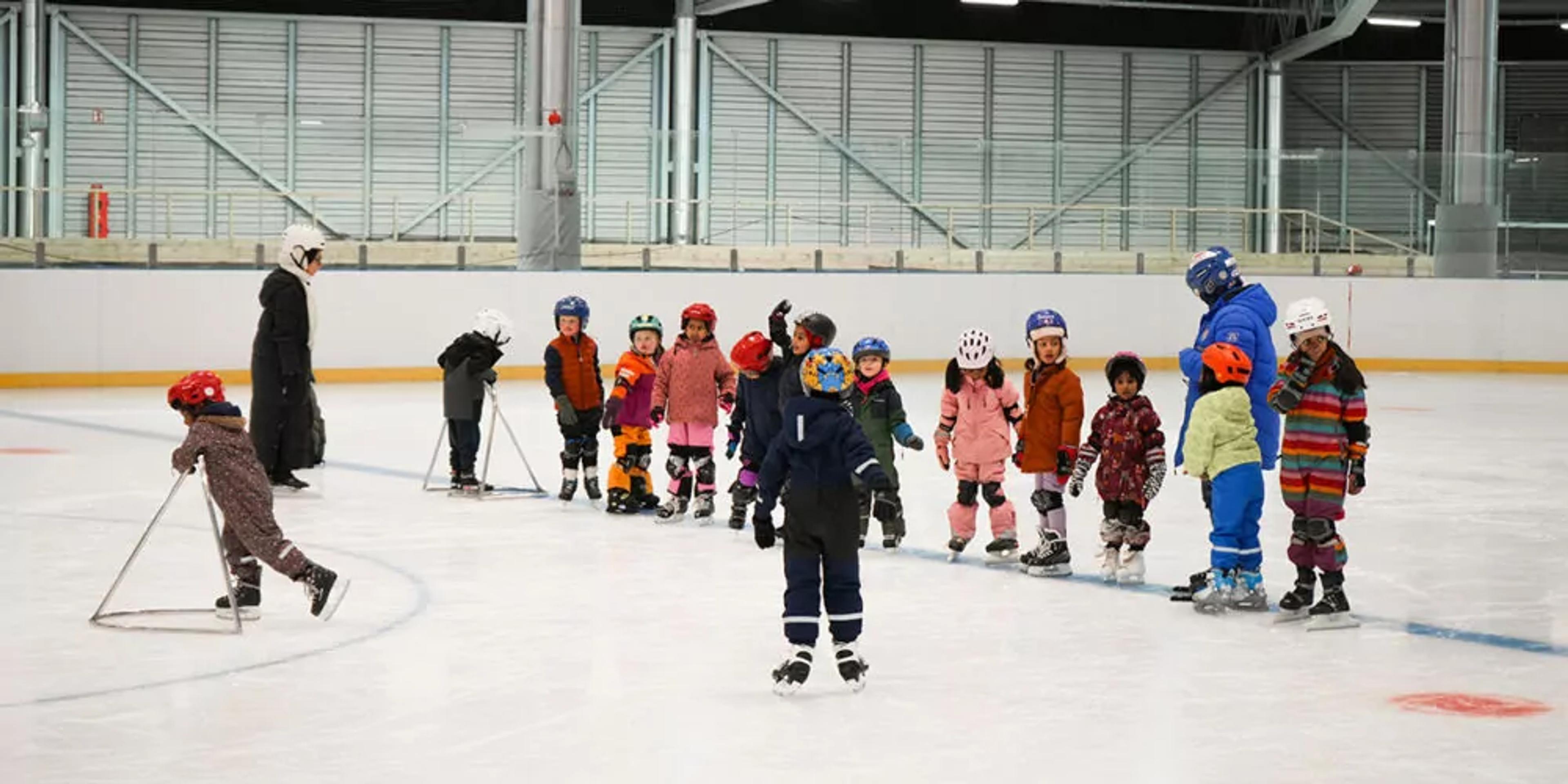 En grupp barn i färgglada hjälmar och skridskor står på isen i en ishall, medan en vuxen hjälper till med övningarna under en skridskoskola. Några barn använder stödramar för att hålla balansen.