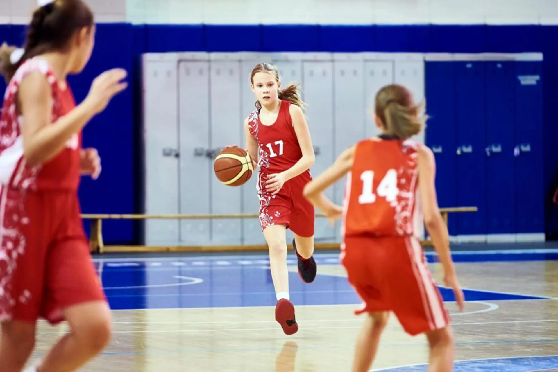 Tre barn i röda basketkläder spelar match i en gympasal, där en flicka med nummer 17 dribblar bollen framåt på planen.