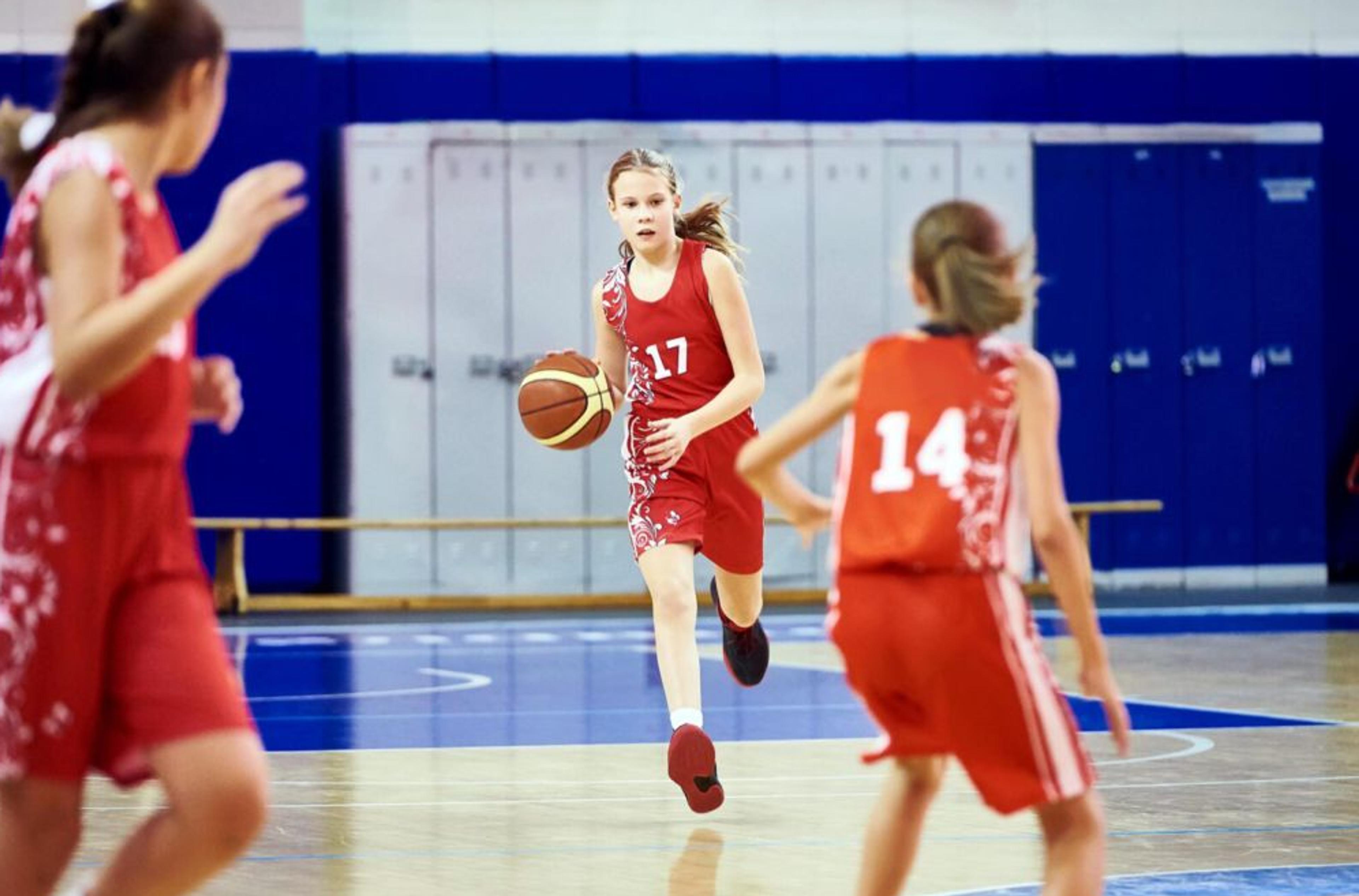 Tre barn i röda basketkläder spelar match i en gympasal, där en flicka med nummer 17 dribblar bollen framåt på planen.
