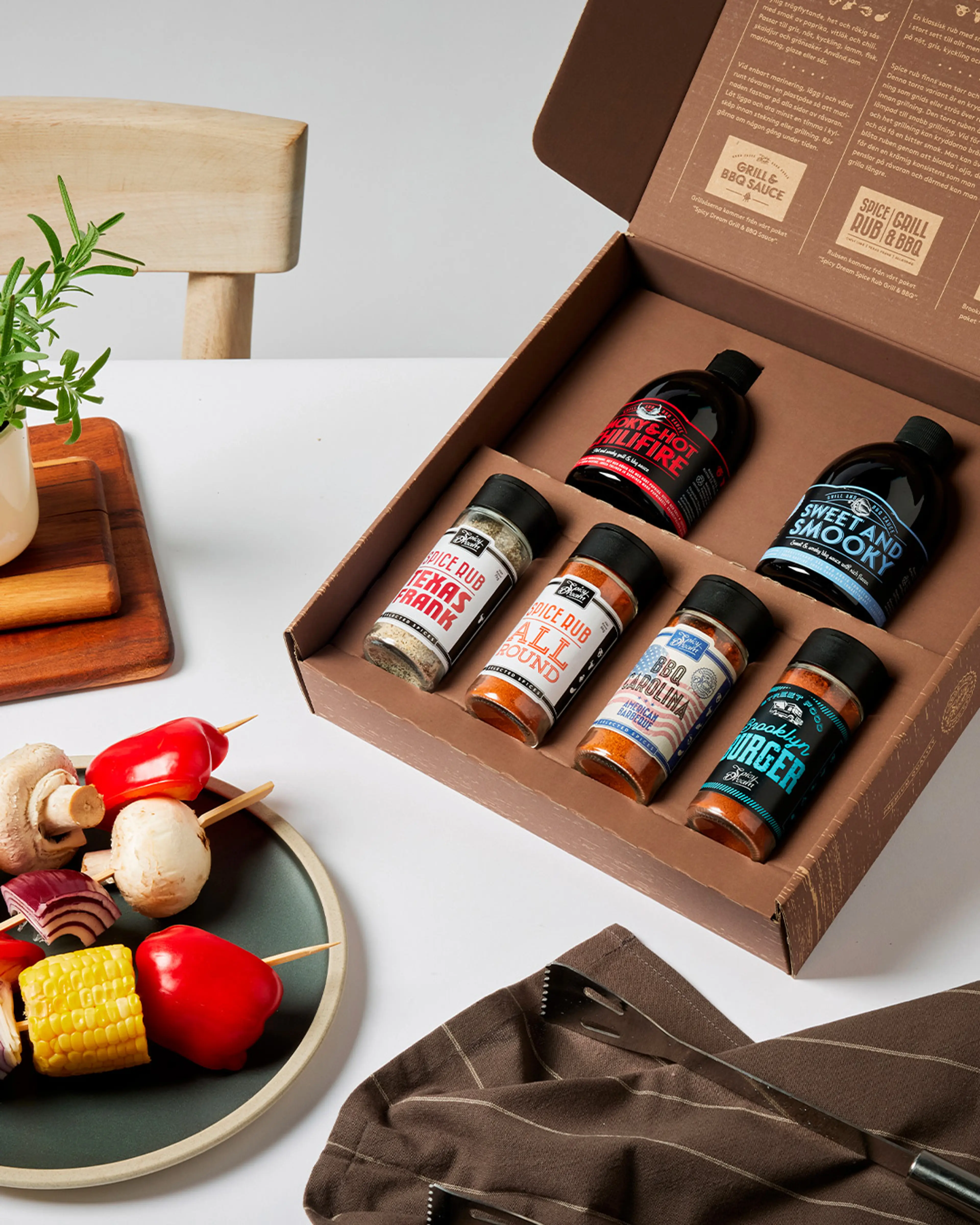 En öppen låda med fem kryddburkar och två flaskor med grillsåser i olika smaker, placerad på ett vitt bord bredvid en tallrik med grillspett av grönsaker och en brun kökshandduk.