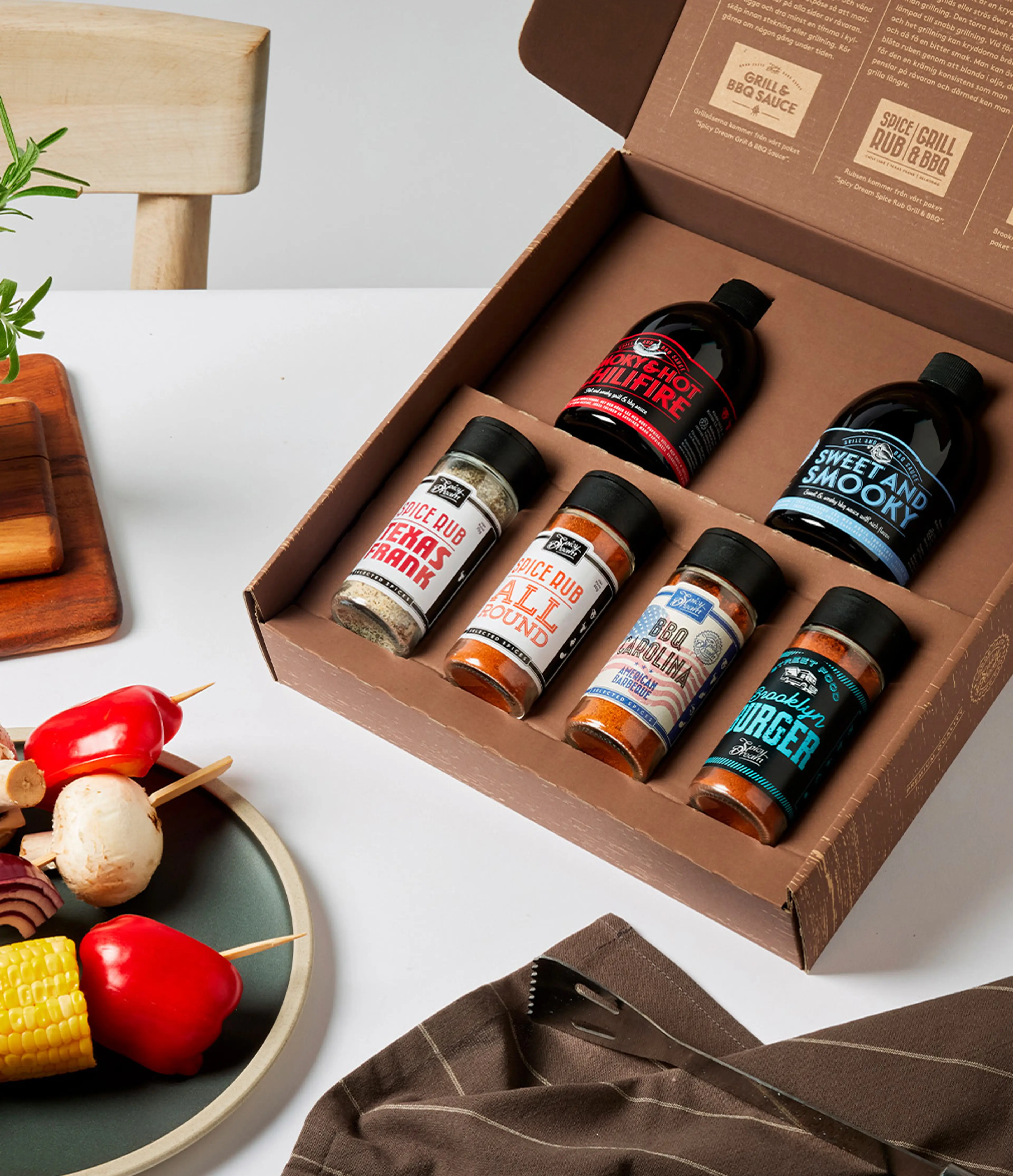 En öppen låda med fem kryddburkar och två flaskor med grillsåser i olika smaker, placerad på ett vitt bord bredvid en tallrik med grillspett av grönsaker och en brun kökshandduk.