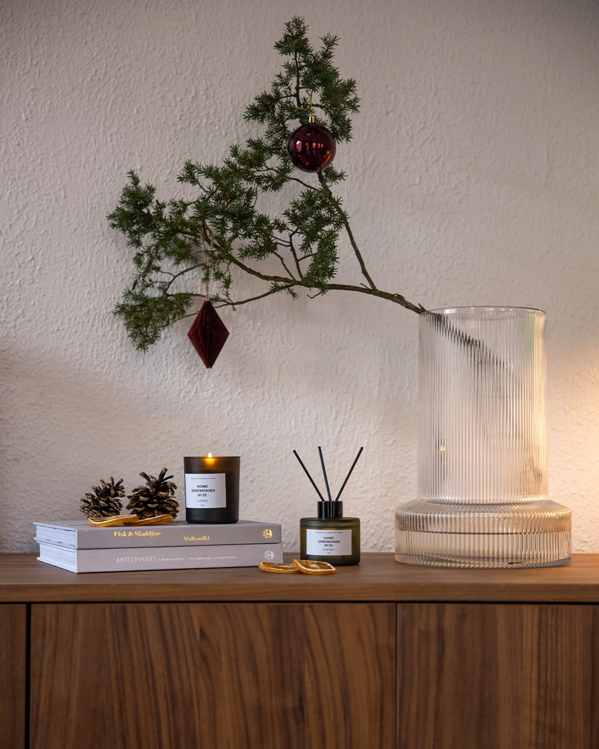 En enkel juldekoration med en kvist i en klar glasvas, prydd med röda julgranskulor. Bredvid står ett doftljus och en doftpinne i mörkgrönt glas, placerade på en träbänk tillsammans med torkade ap
