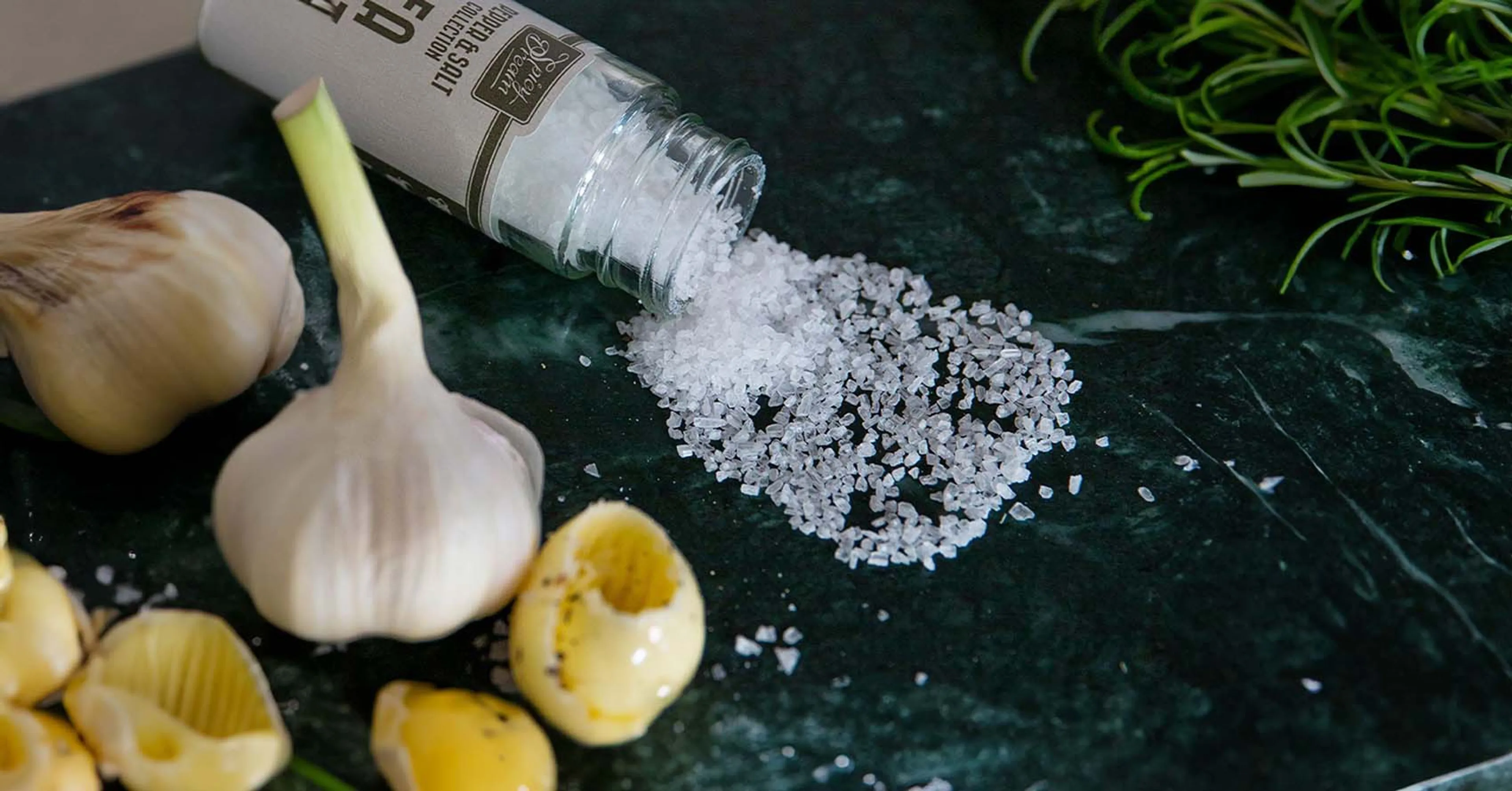Ett glasrör med grovt havssalt som är urtappat på en mörk grön marmorbänk. I bakgrunden syns en vitlök, vitlöksskivor och en kvist rosmarin.