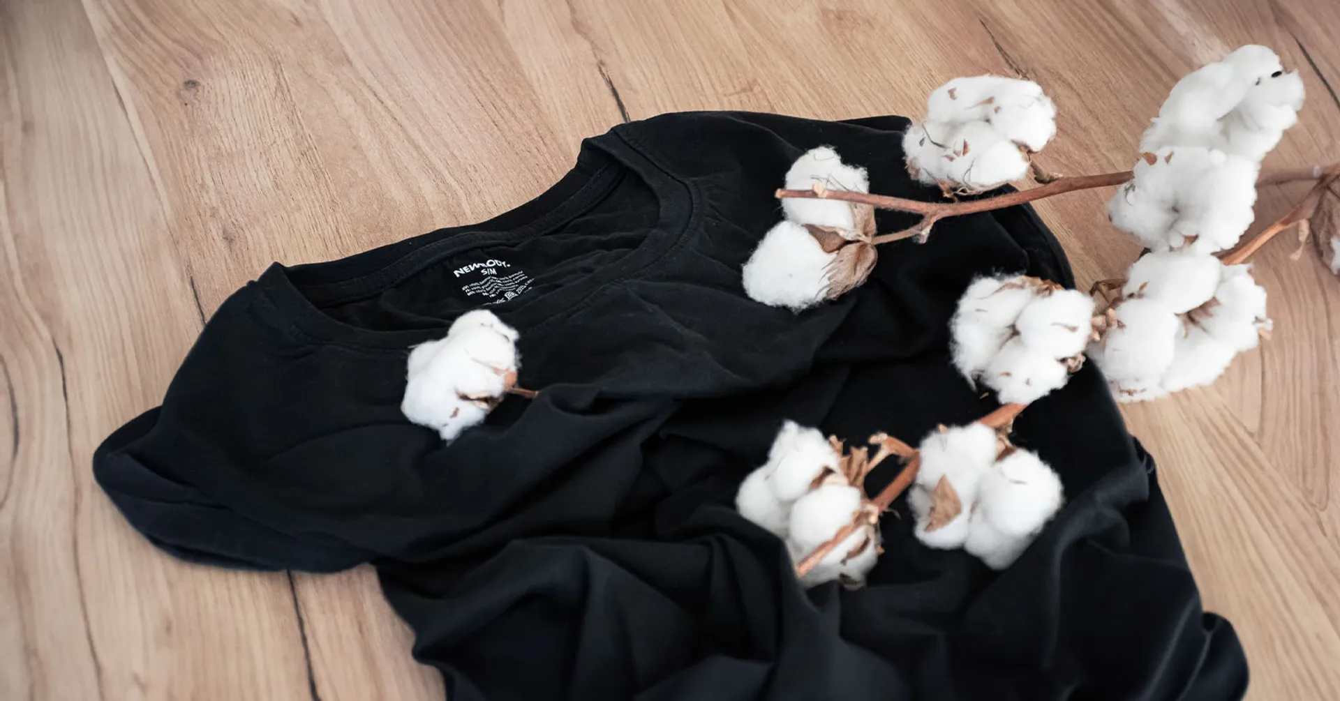 Svart, mjuk t-shirt i bomull ligger tillsammans med några bomullskvistar på ett ljust träunderlag.