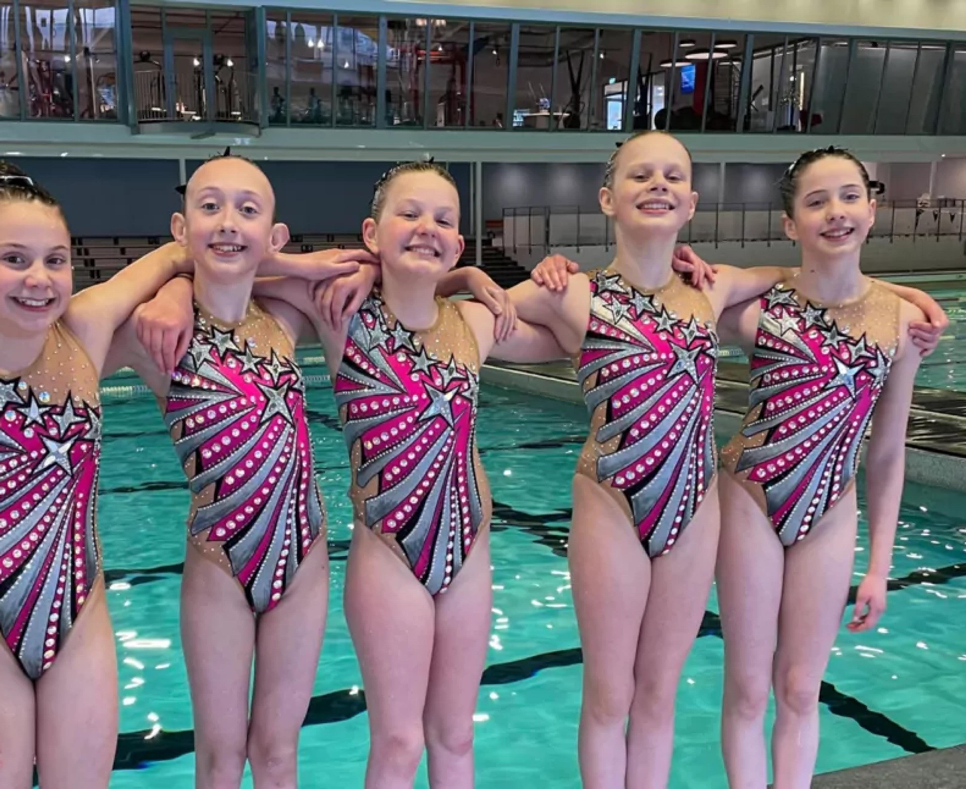 Fem unga simmare i färgglada, mönstrade dräkter står nära varandra vid poolkanten med armarna runt varandra. Bakgrunden visar en simhall med tomma läktare och bassäng.