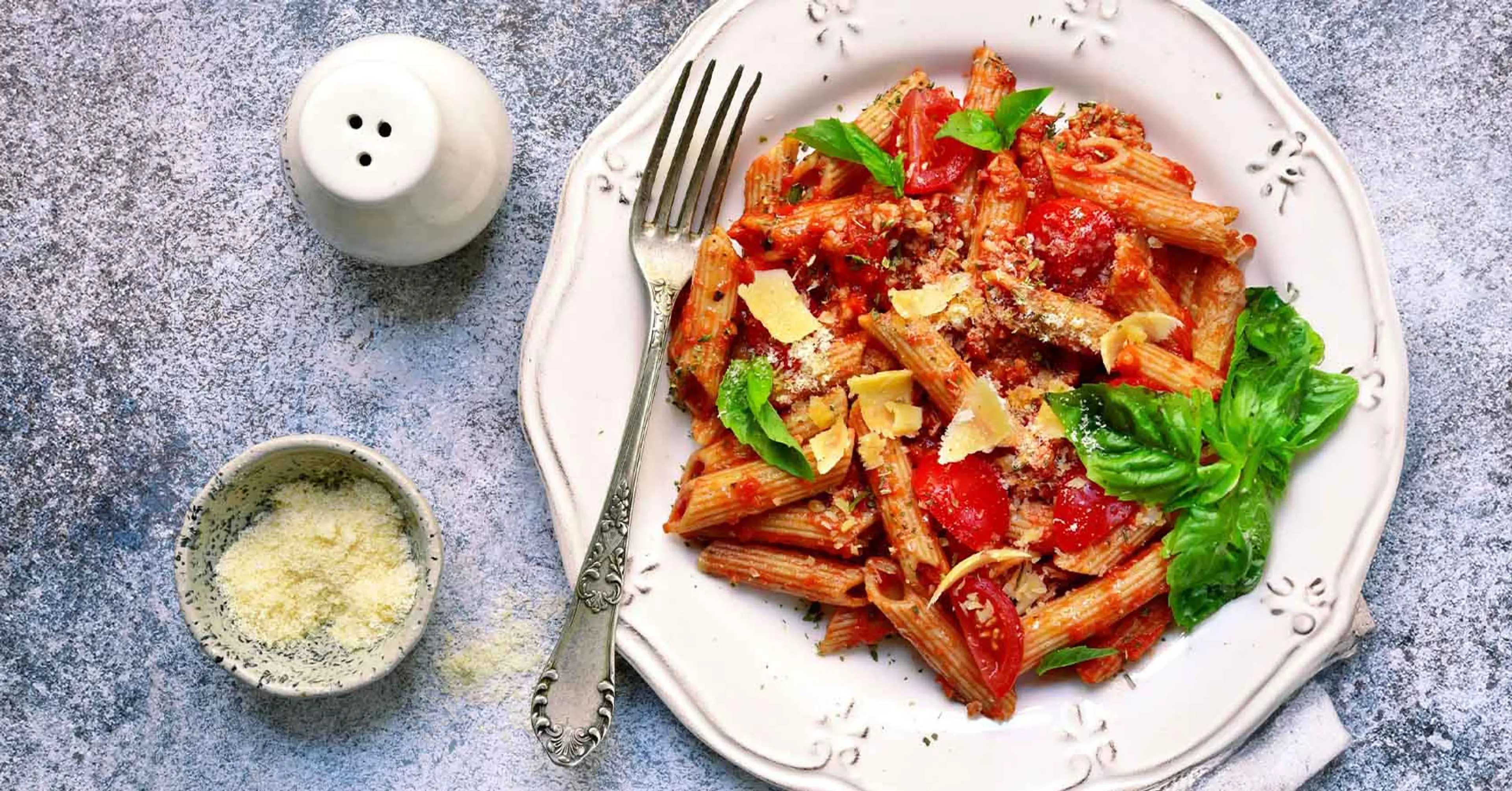 En tallrik med pennepasta blandad med tomatsås, körsbärstomater, basilika och riven ost. Bredvid ligger en liten skål med mer riven ost och en vit saltkar.
