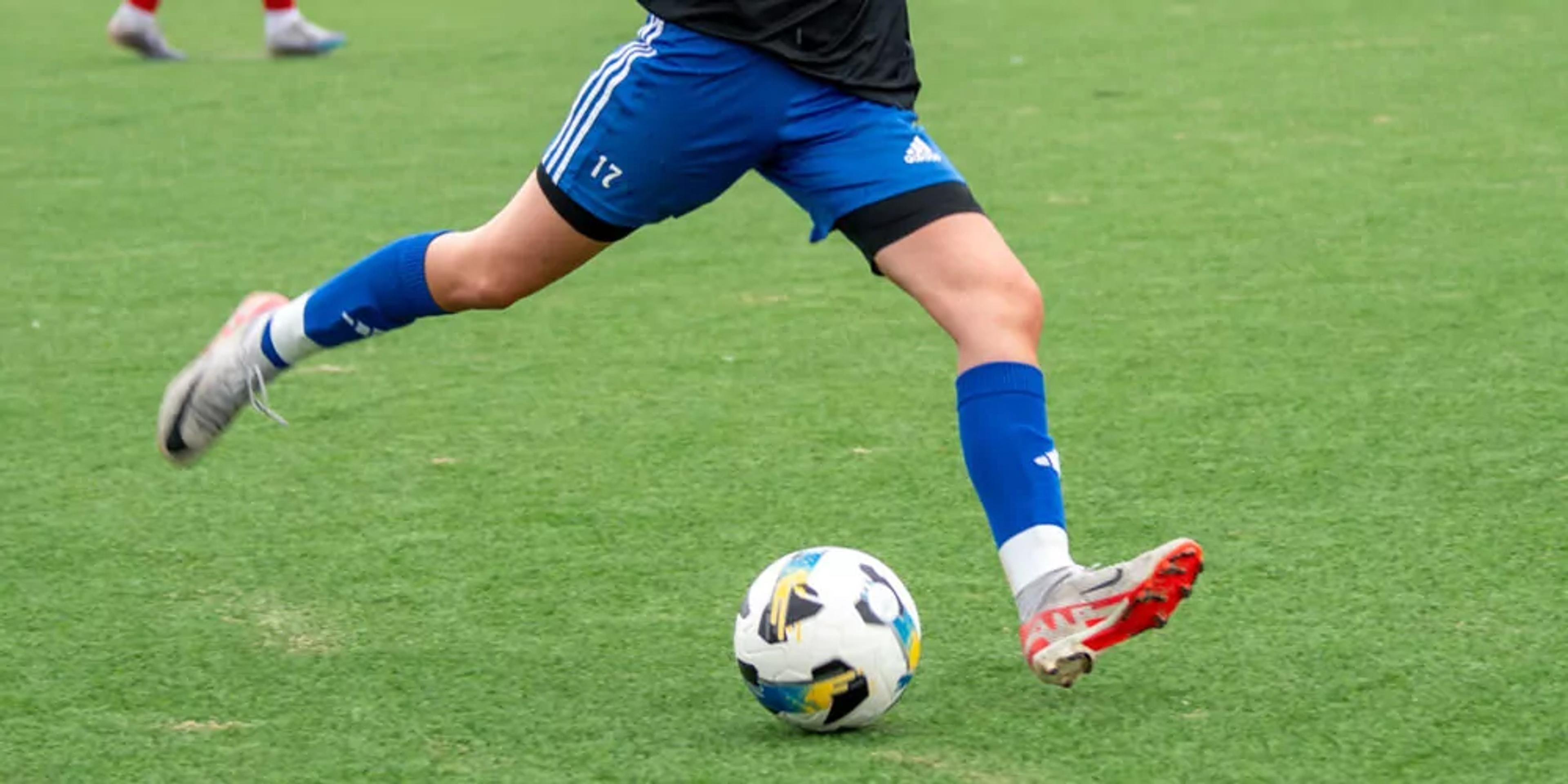En person i blå fotbollsshorts och blå strumpor sparkar en vit fotboll med färgglada mönster på en grön gräsplan. Endast benen syns i rörelse.