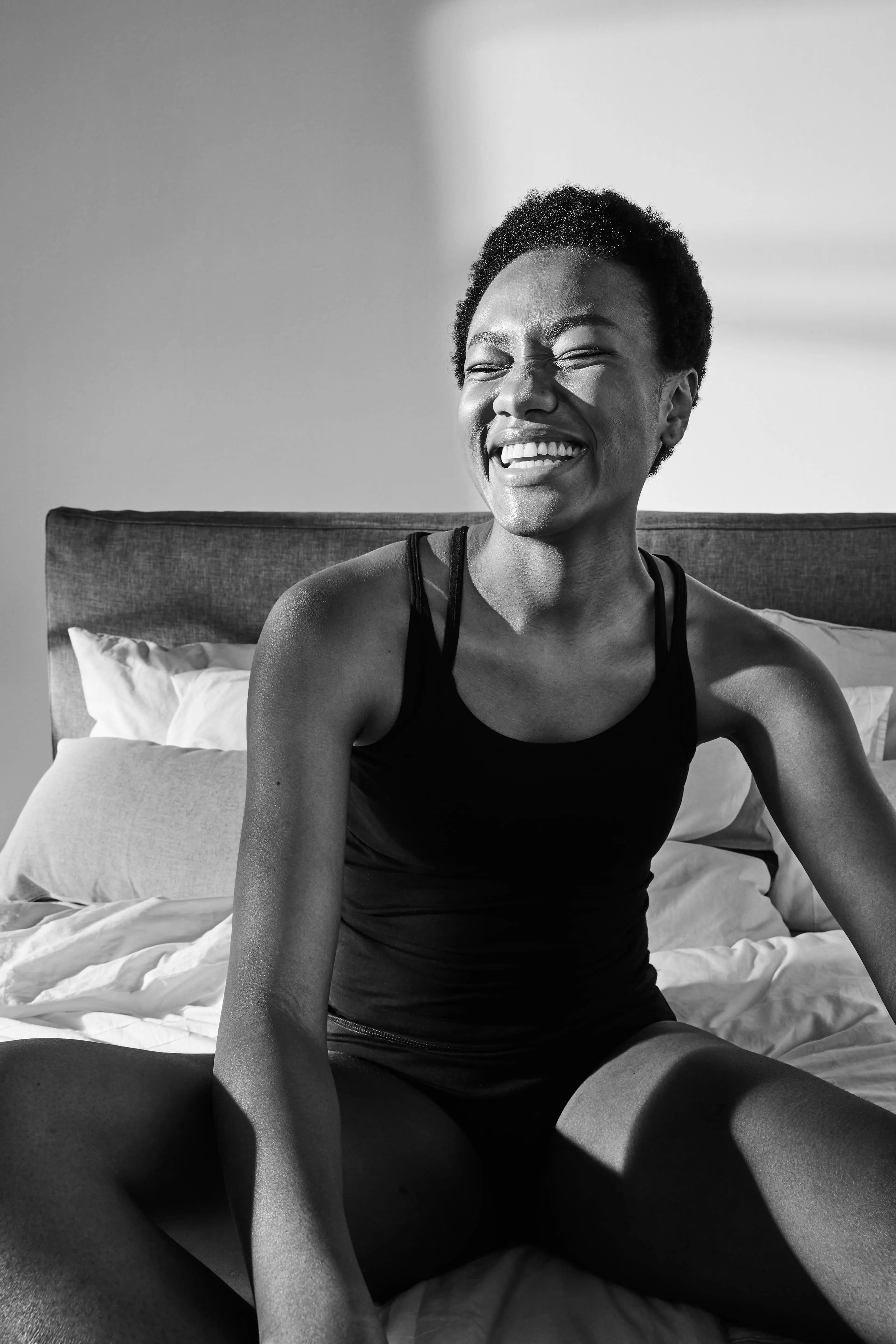 En person i svart linne och underkläder sitter avslappnat i en säng med vitt sänglinne. Solen kastar mjuka skuggor över kroppen och väggarna i rummet.