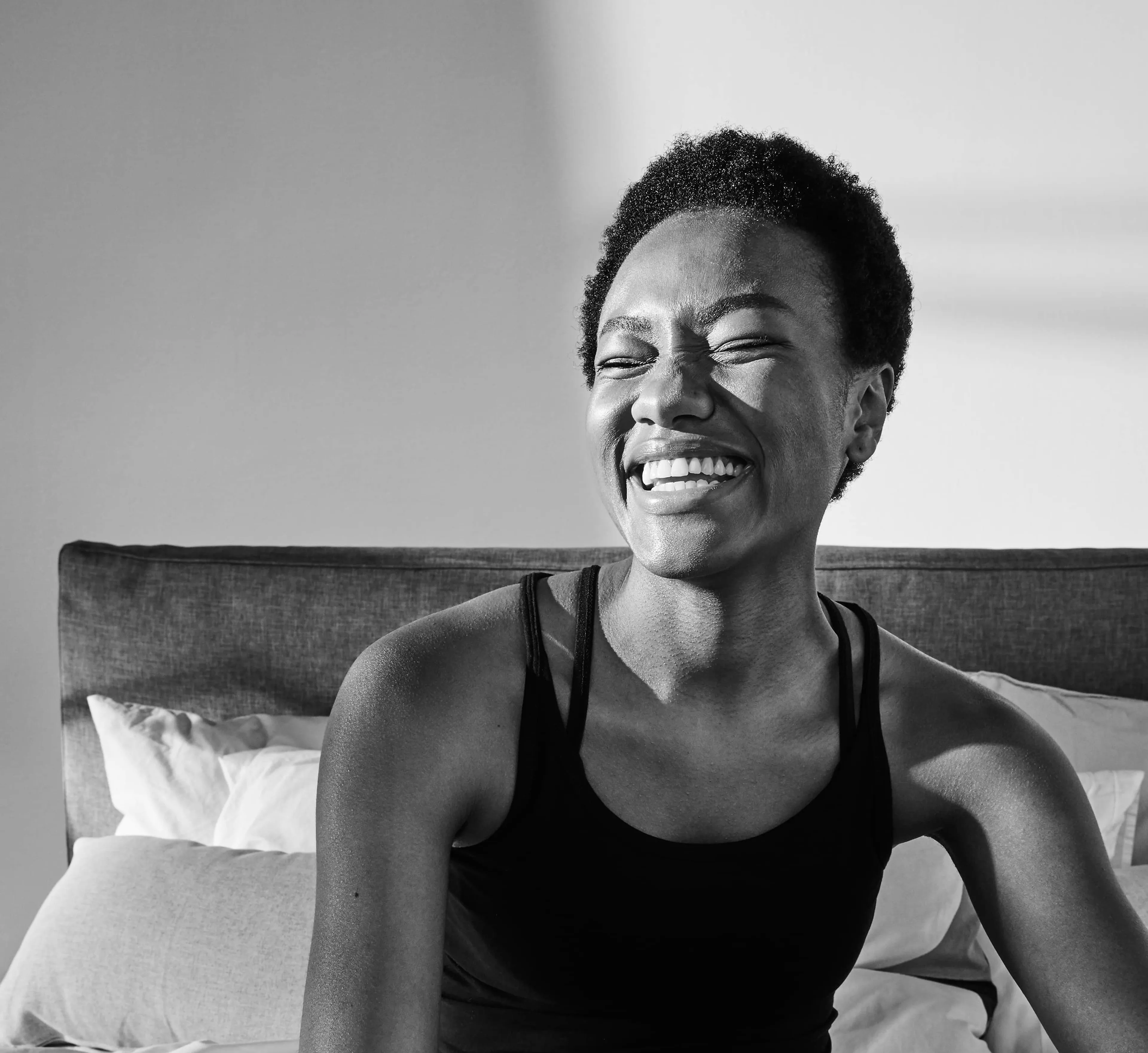 En person i svart linne och underkläder sitter avslappnat i en säng med vitt sänglinne. Solen kastar mjuka skuggor över kroppen och väggarna i rummet.